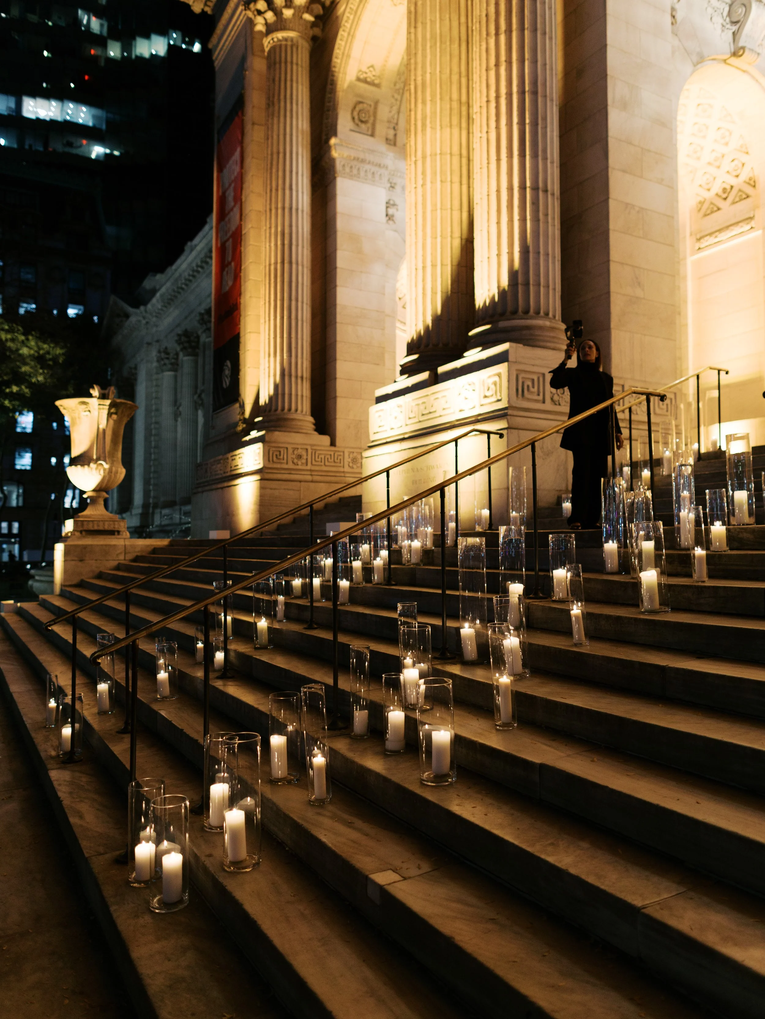 Nighttime scene of candles in glass containers lined along the steps leading up to a grand, illuminated building with large columns. A person is standing on the steps, taking a photo or video with a smartphone.