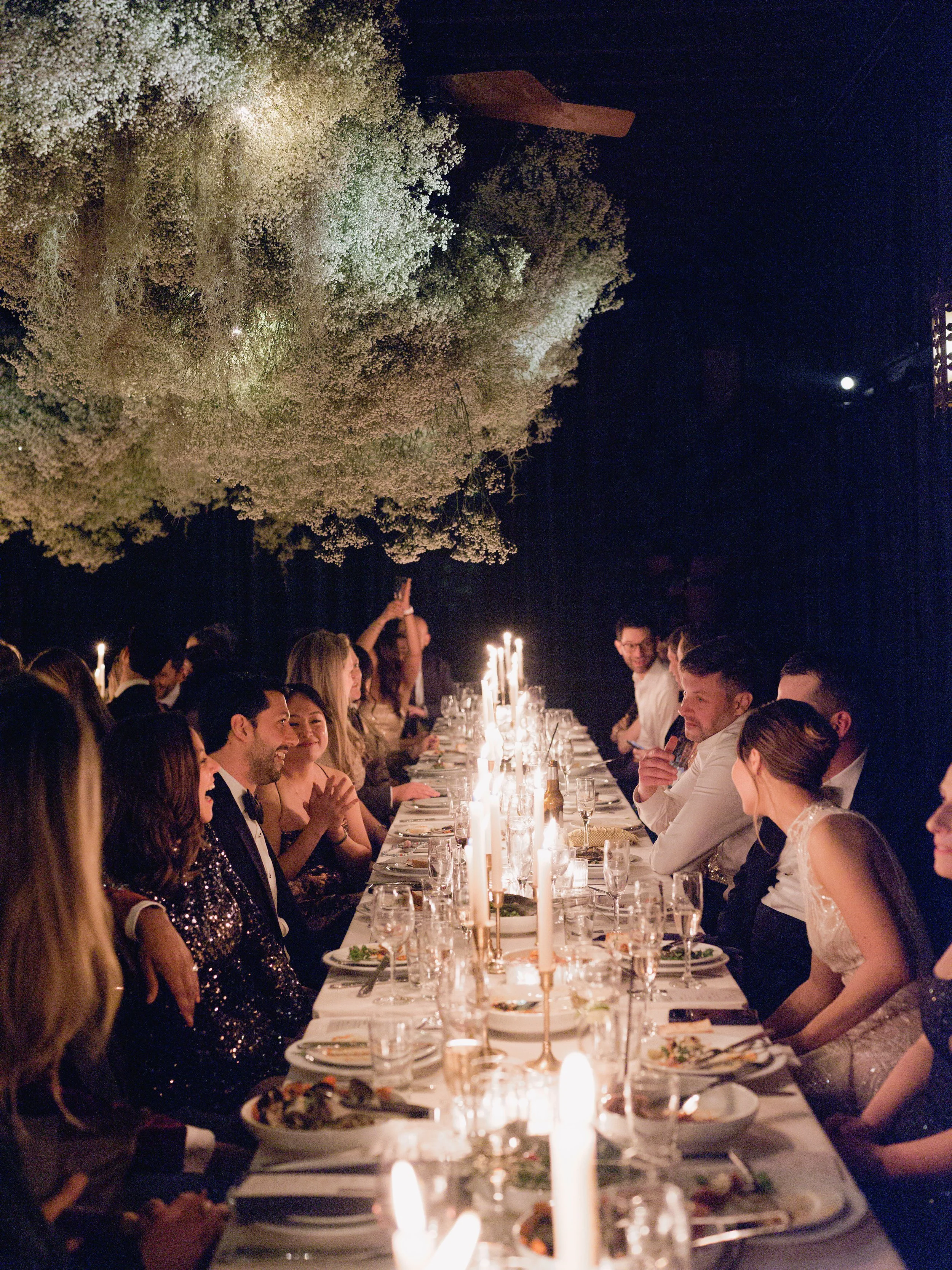 A formal dinner party with guests seated at a long table decorated with candles, plates, and glassware, underneath hanging flowers at night.