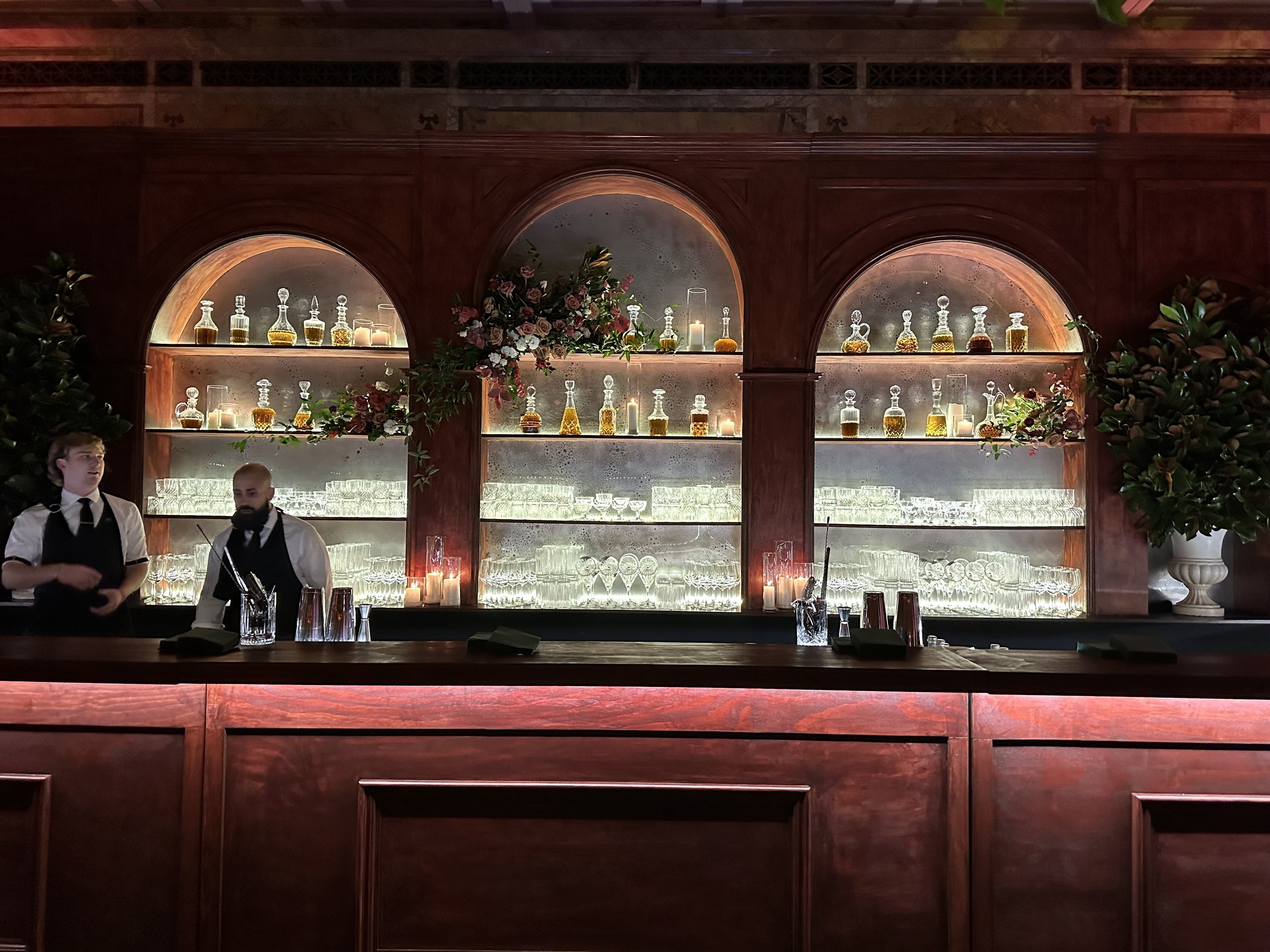 Bar with wooden shelves displaying bottles of alcohol, glassware, and candles, with two bartenders standing behind the counter.