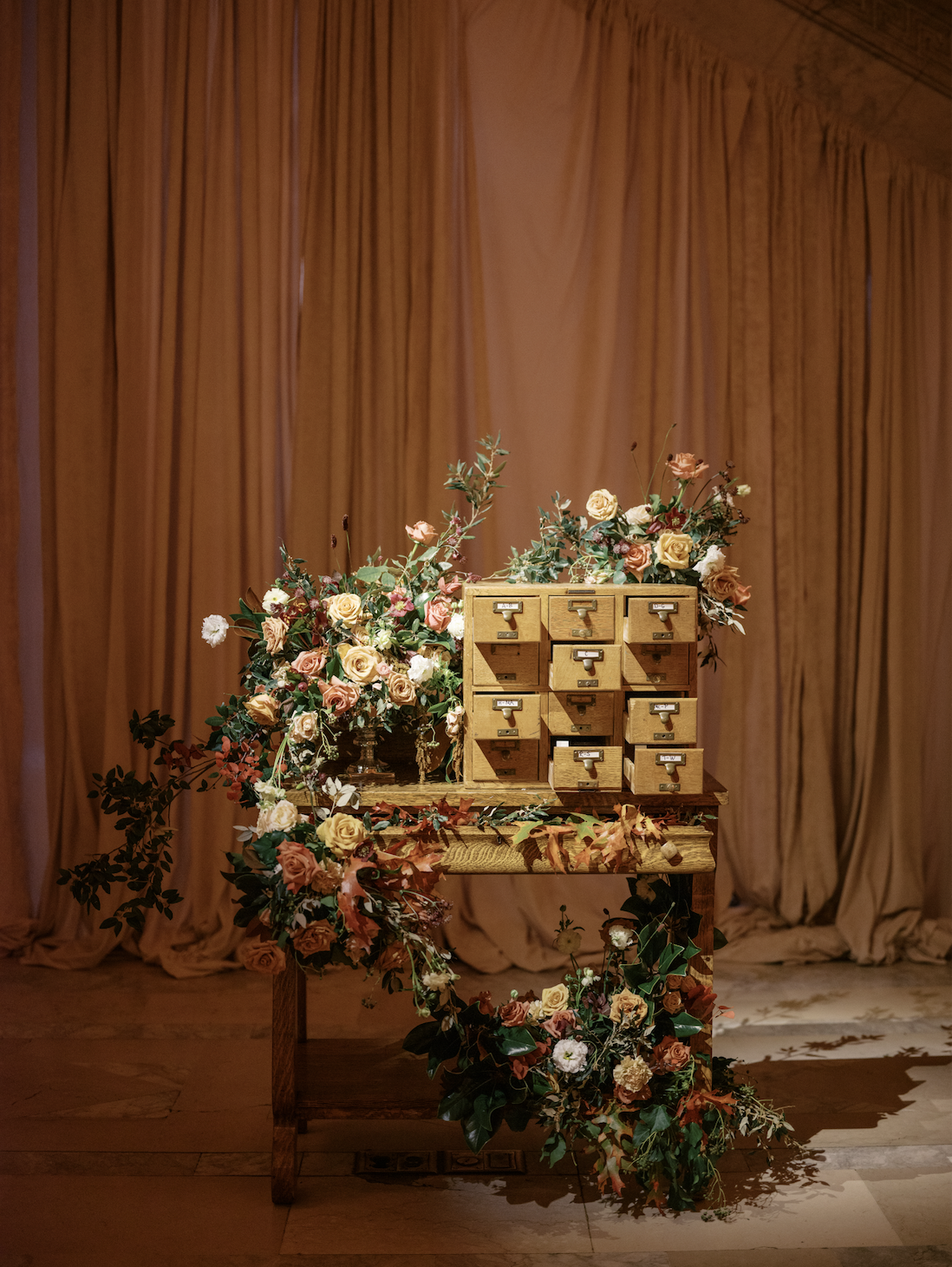 Wooden desk with small drawers over floral arrangements of roses and carnations, with a pink curtain backdrop.