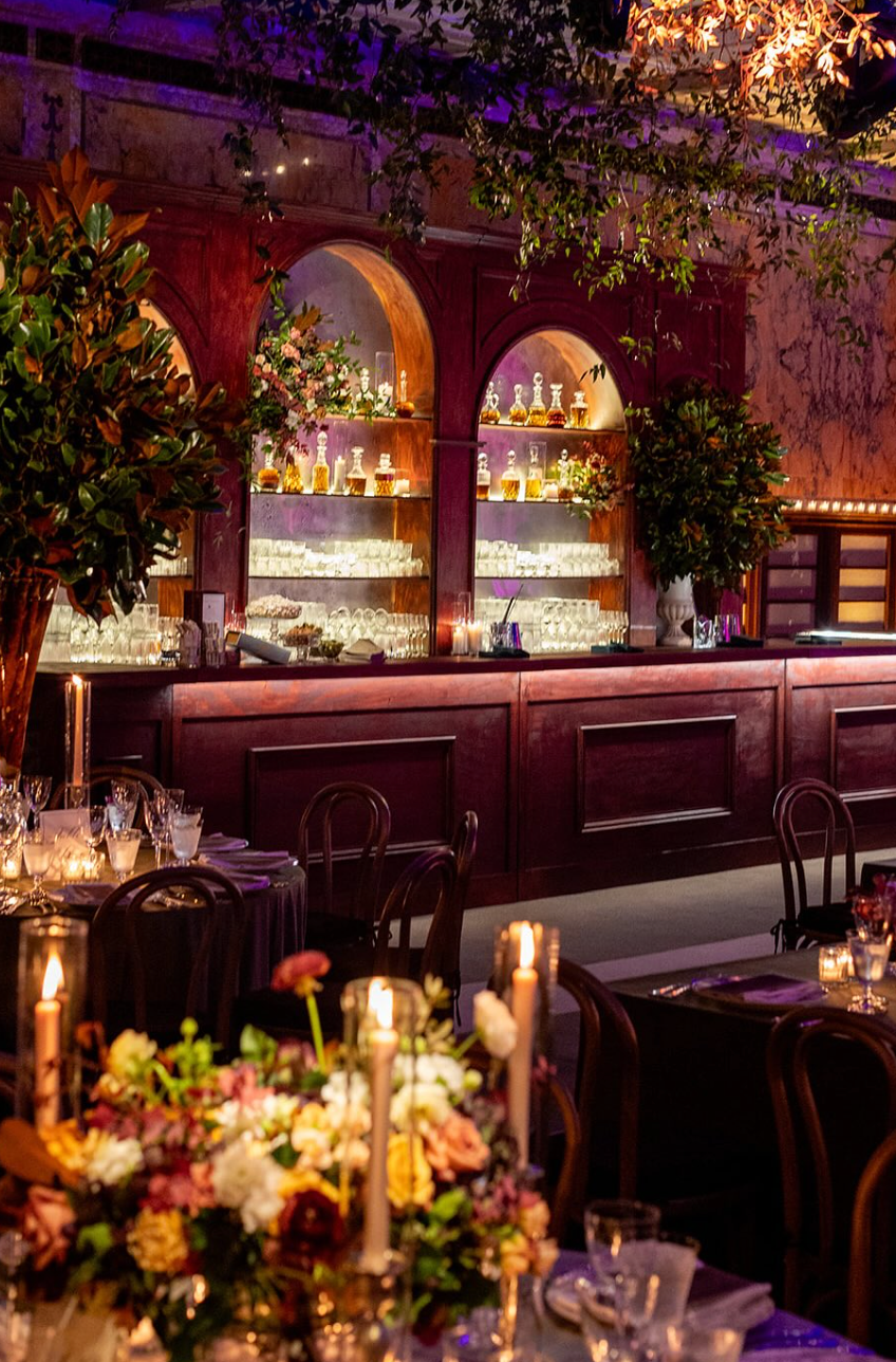 Elegant restaurant bar with a polished wooden counter, illuminated glass shelves with bottles and glassware, floral arrangements, and lit candles on tables.
