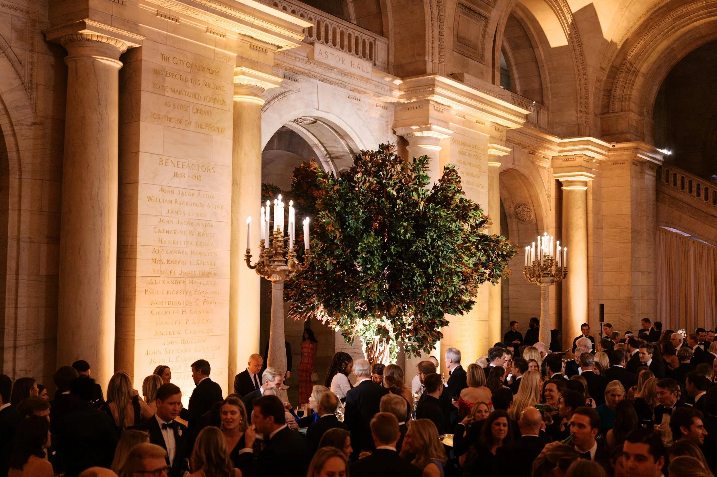 Elegant event inside a grand hall with classical architecture, tall marble columns, and ornate chandeliers. A large tree centerpiece with candles surrounds a crowd of formally dressed people.