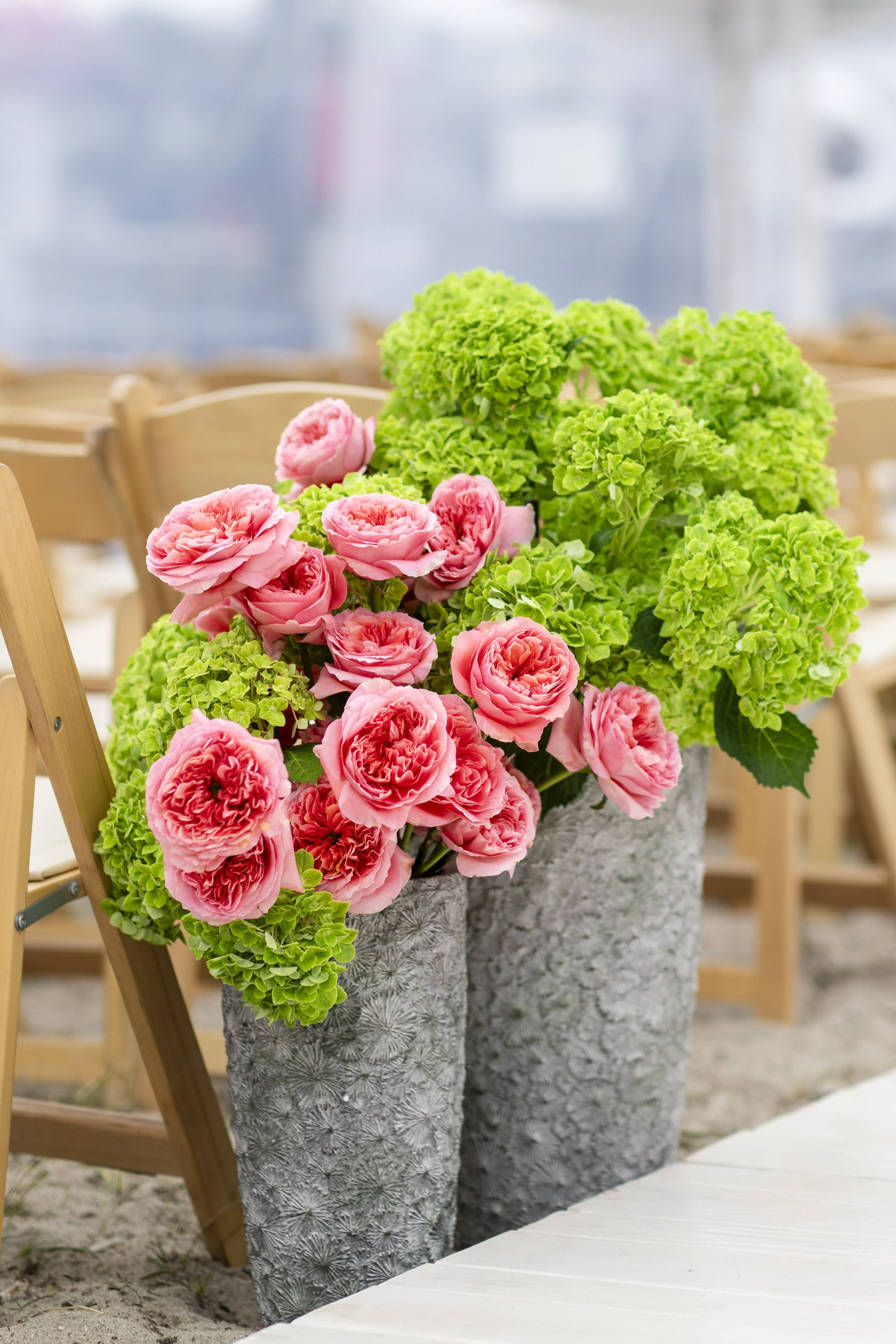 Pink ranunculus flowers and green hydrangeas in textured gray vases at an outdoor event.