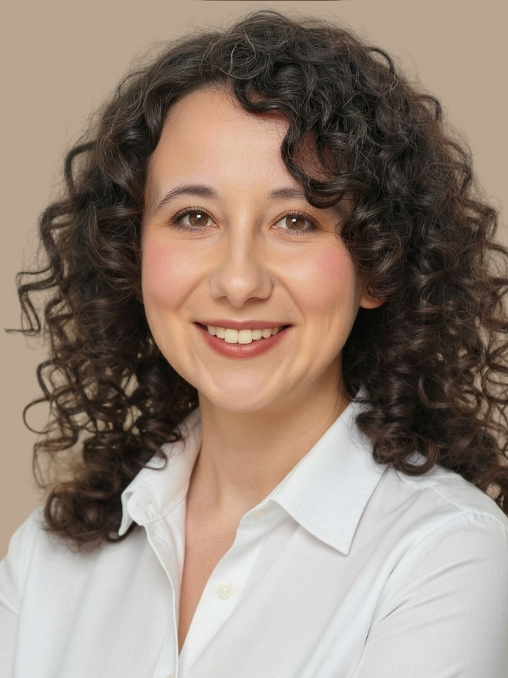 A woman with curly dark hair and fair skin smiling in a white collared shirt against a beige background.