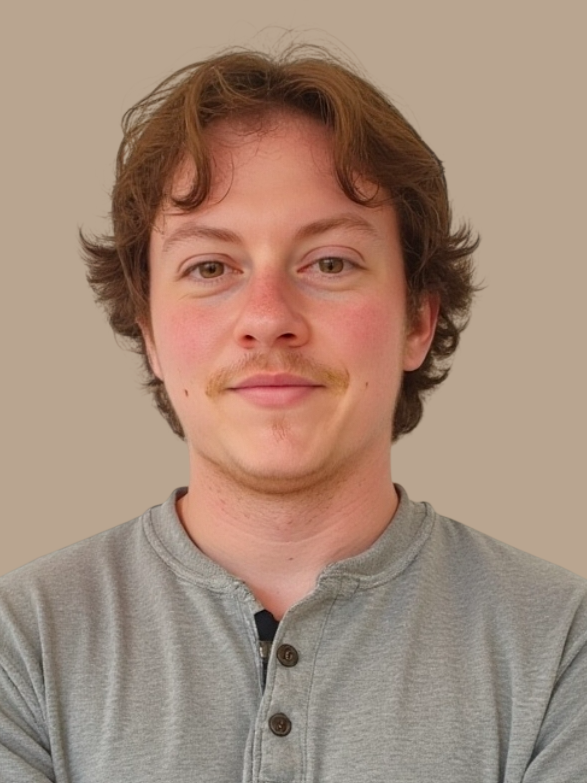 Portrait of a young man with light brown hair, slight facial hair, and a neutral expression, wearing a gray henley shirt, standing against a plain beige background.