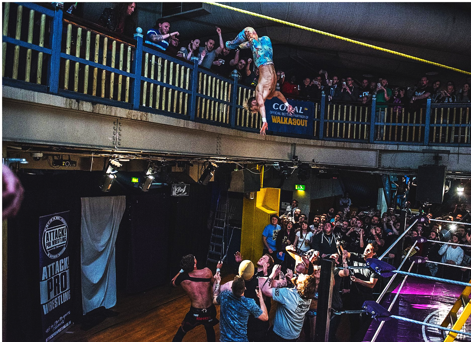 A professional wrestling event with a wrestler performing a backflip from the top rope in the ring, while the crowd watches and films. The venue has two levels of spectators and banners, including one for Attack Pro Wrestling.