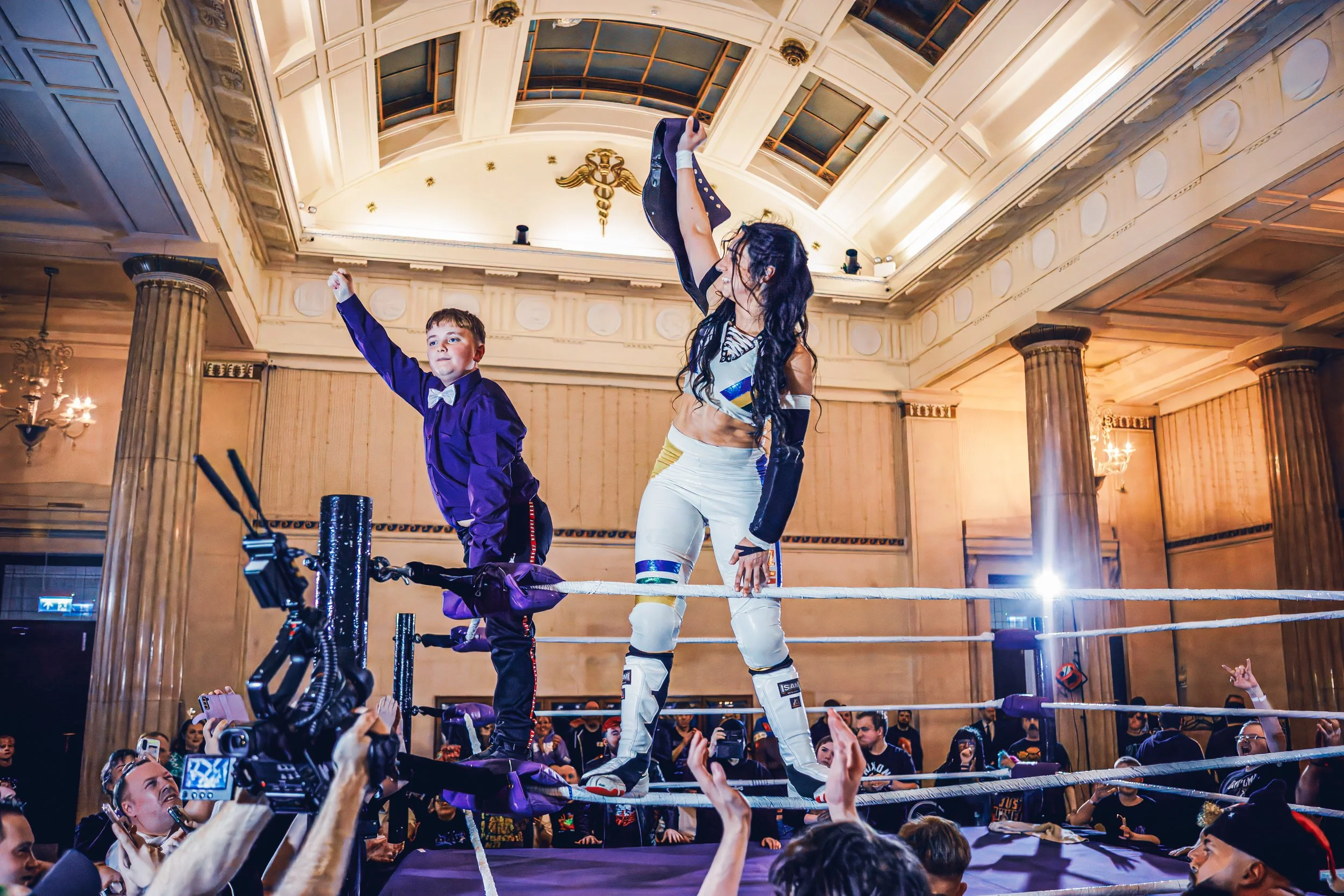 A professional wrestling match with a young boy and a woman standing on the turnbuckle in a large, ornate hall filled with spectators.