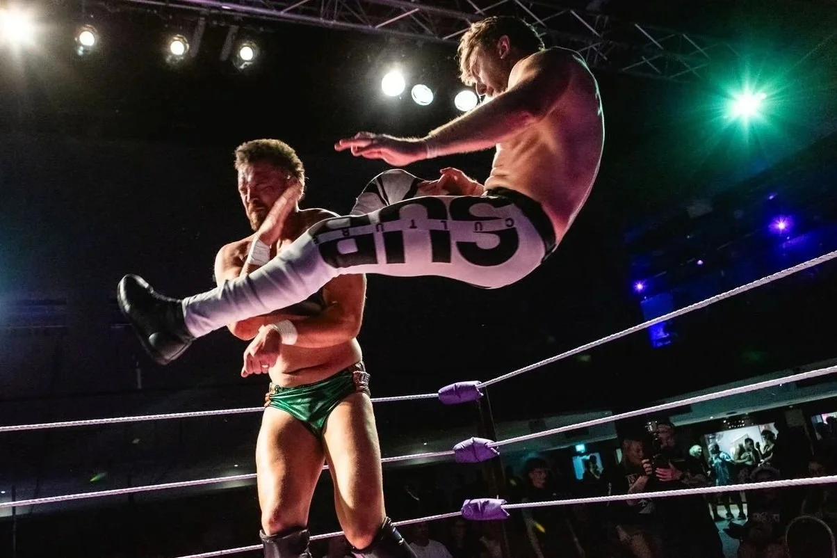A professional wrestling event with a wrestler performing a backflip from the top rope in the ring, while the crowd watches and films. The venue has two levels of spectators and banners, including one for Attack Pro Wrestling.