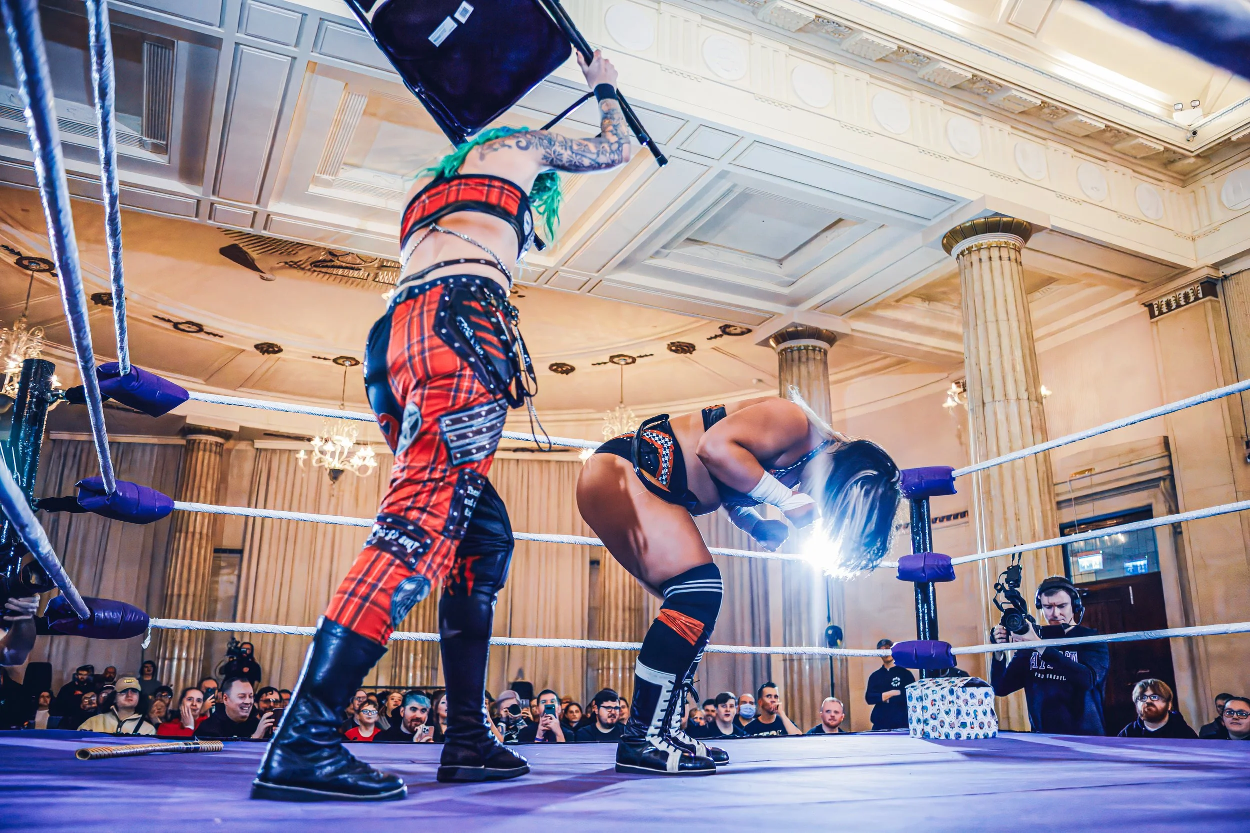 Two female wrestlers in a wrestling ring during a match, with an audience watching and a cameraman recording near the ring, in an ornate indoor venue.