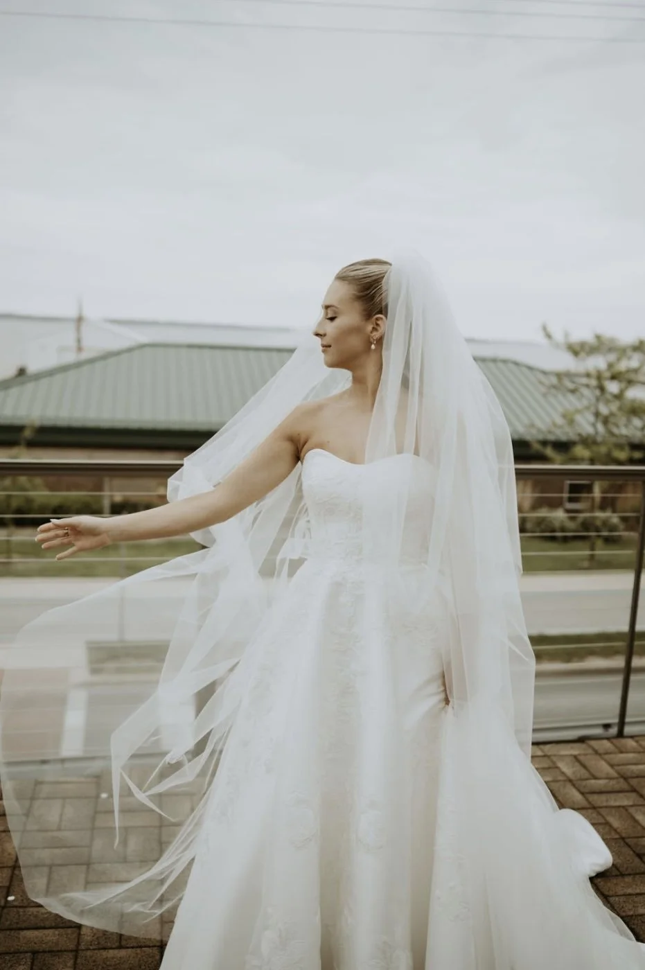 A bride in a strapless white wedding gown and veil standing outdoors on a balcony, with her eyes closed and one arm outstretched.