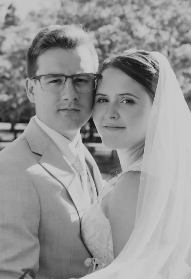 A black and white photo of a bride and groom close together, outdoors, with trees and a fence in the background.