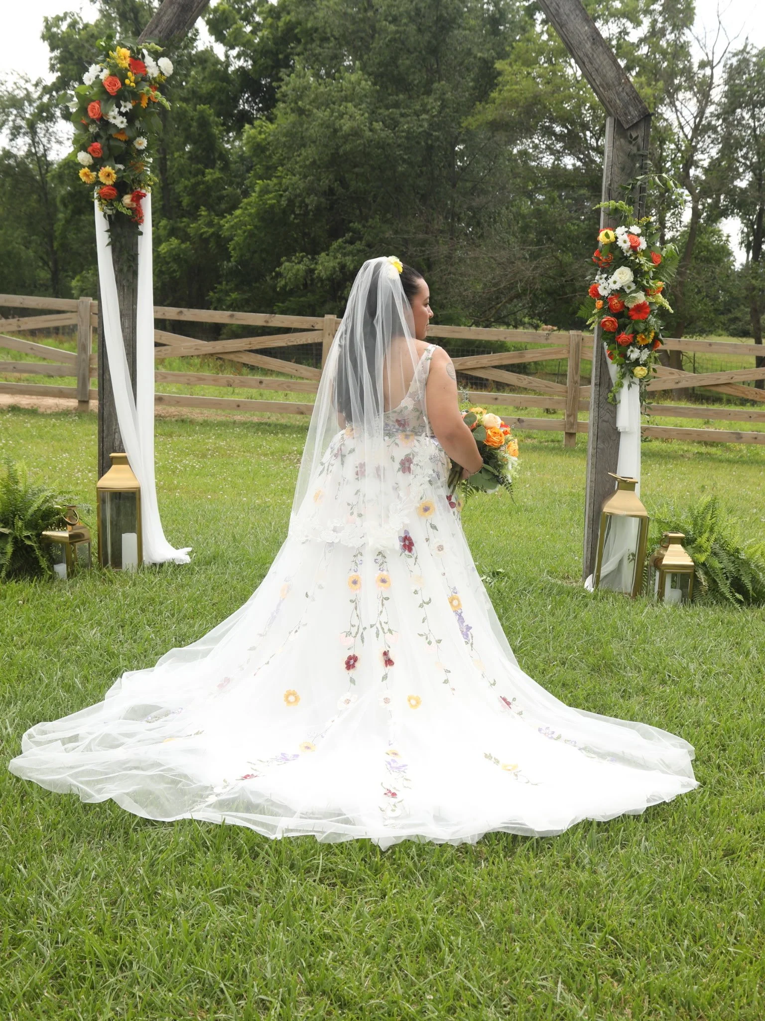A bride in a white wedding dress with floral embroidery, holding a bouquet, standing outdoors under a wooden arch decorated with colorful flowers on a grassy area surrounded by trees.
