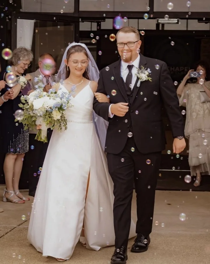 A bride and groom walking arm in arm, smiling, surrounded by floating bubbles, with guests taking photos in the background during their wedding celebration.