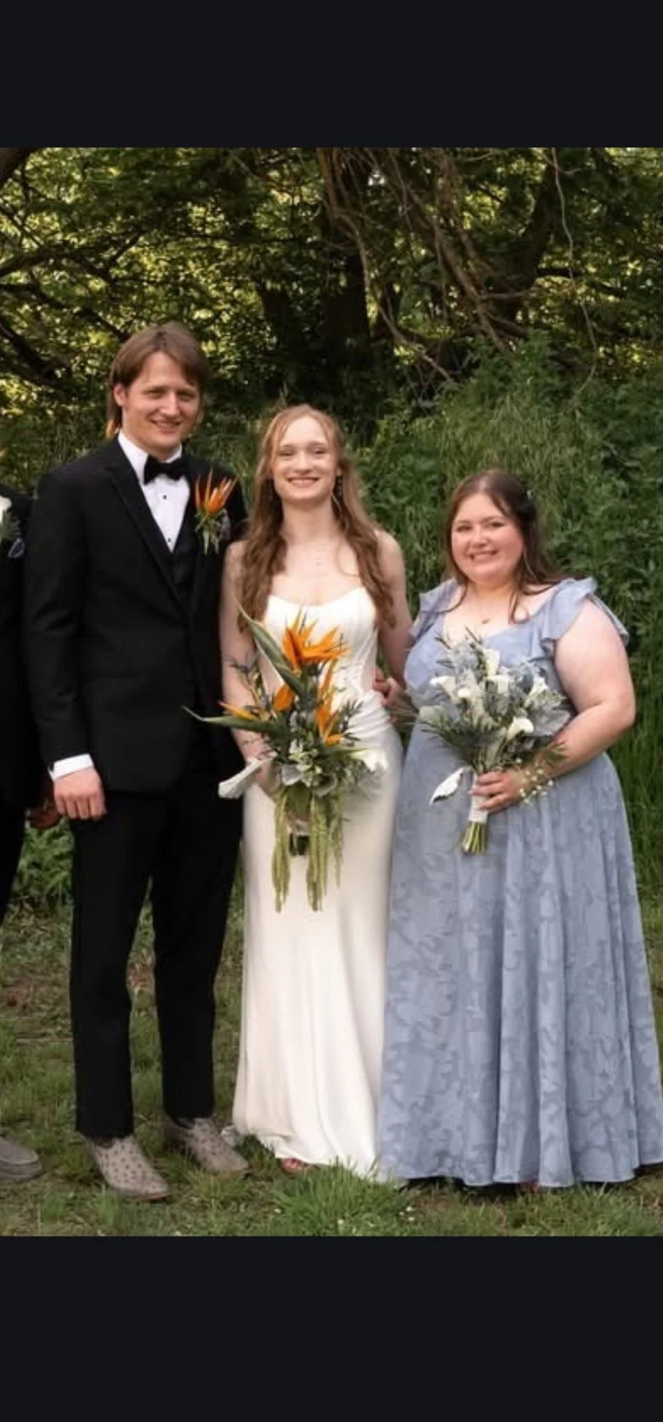 Three people standing outdoors, a man in a tuxedo, a woman in a wedding dress, and a woman in a light blue dress, all holding bouquets of flowers, in front of green trees.