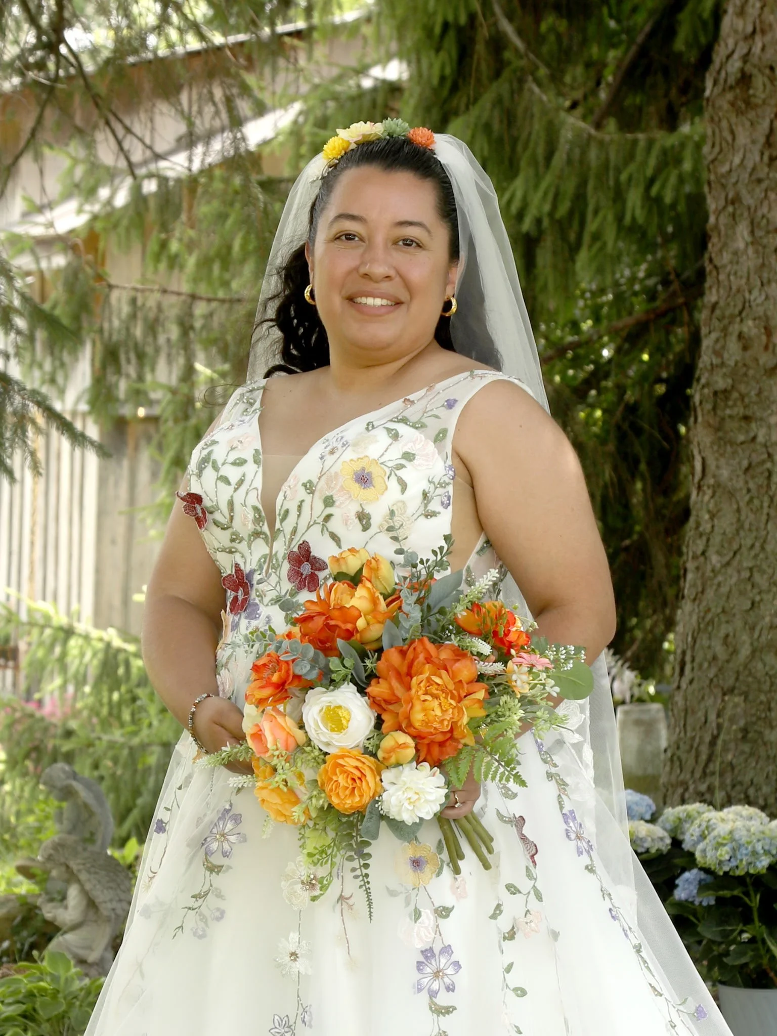 A woman in a floral white dress with a veil and a flower crown holding a colorful bouquet of orange, white, and yellow flowers, standing outdoors with greenery and trees in the background.
