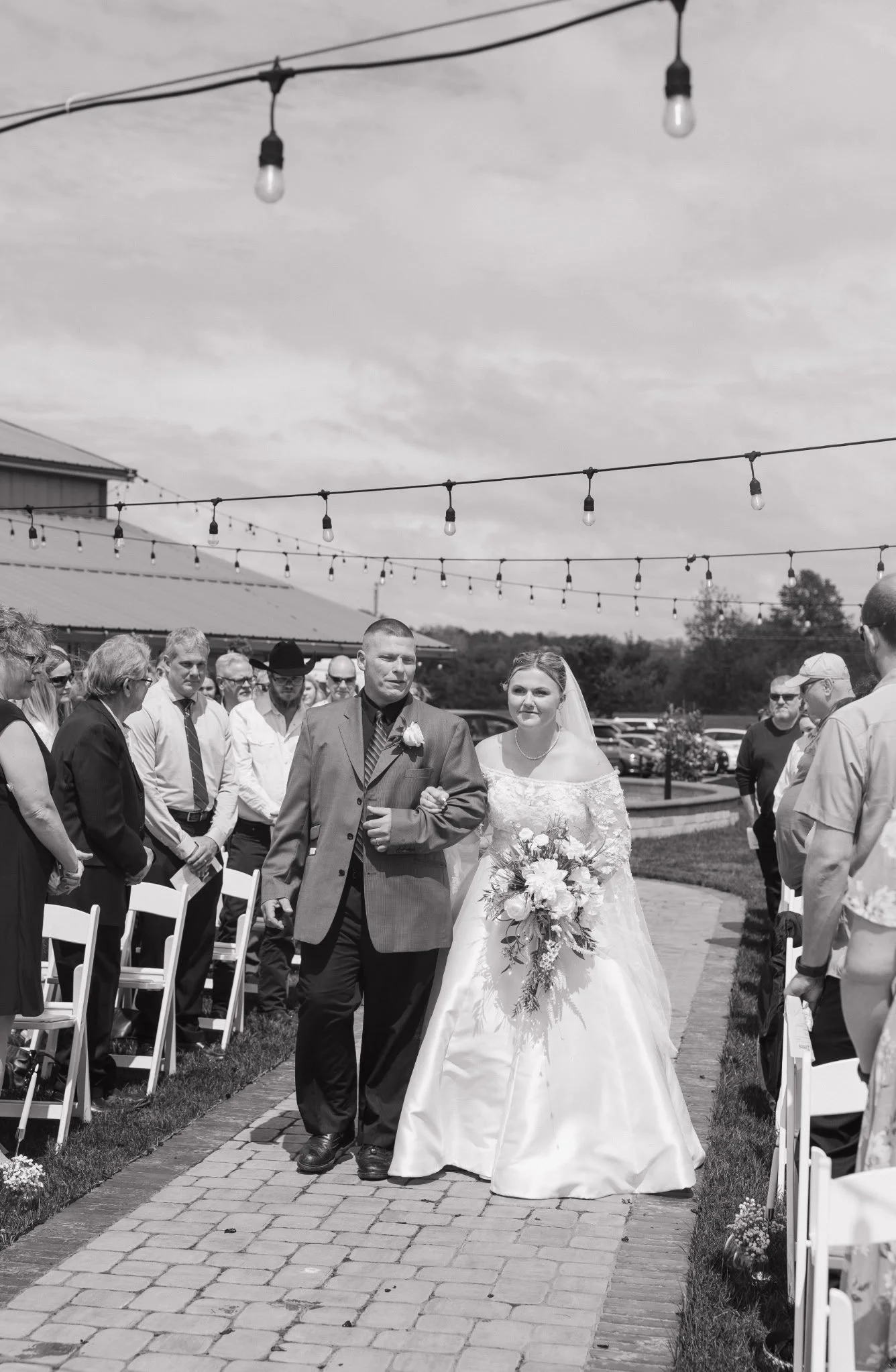 A black and white photograph of a wedding scene. A bride and groom walk down an outdoor aisle lined with white chairs and small flower arrangements, surrounded by guests. Overhead, string lights hang across the sky, and a building is visible in the b