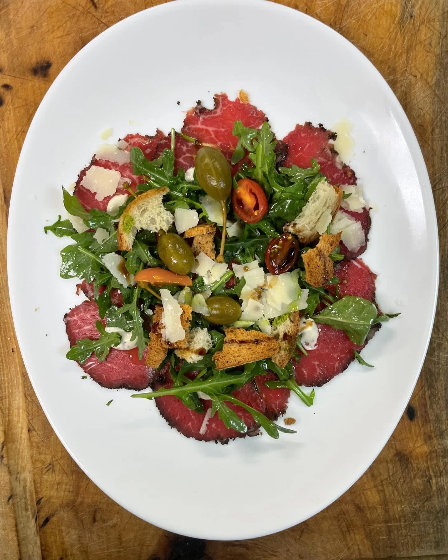 A plate of beef carpaccio garnished with arugula, shaved Parmesan, sliced bread, pickles, cherry peppers, and balsamic glaze.