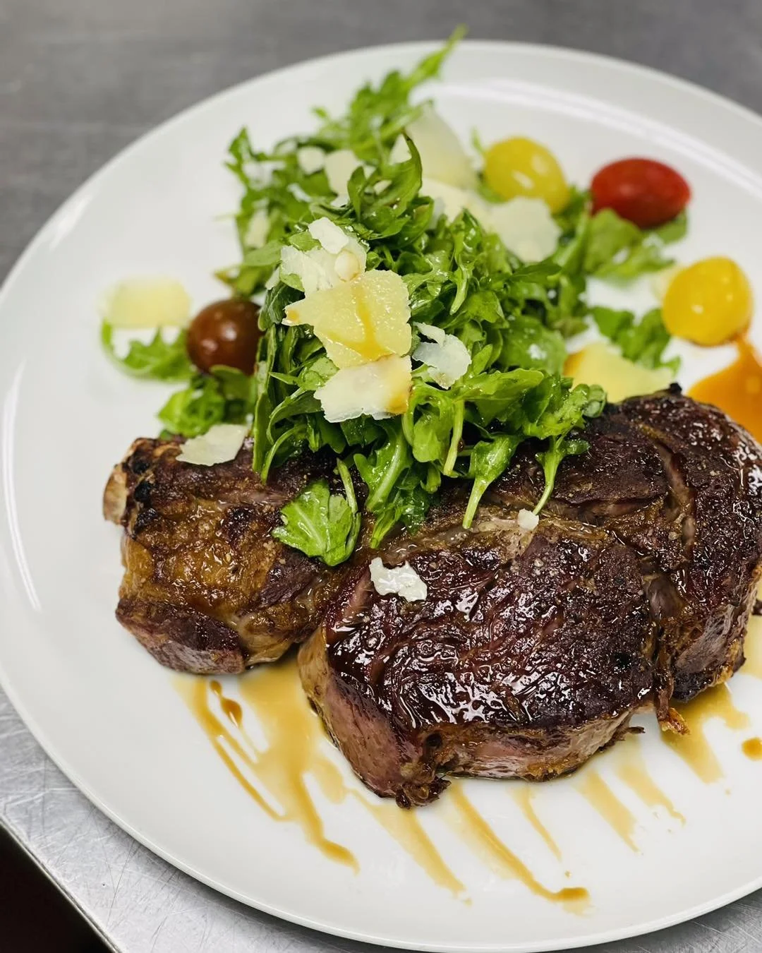 Seared steak topped with arugula salad and shaved Parmesan on a white plate with cherry tomatoes and salad dressing.