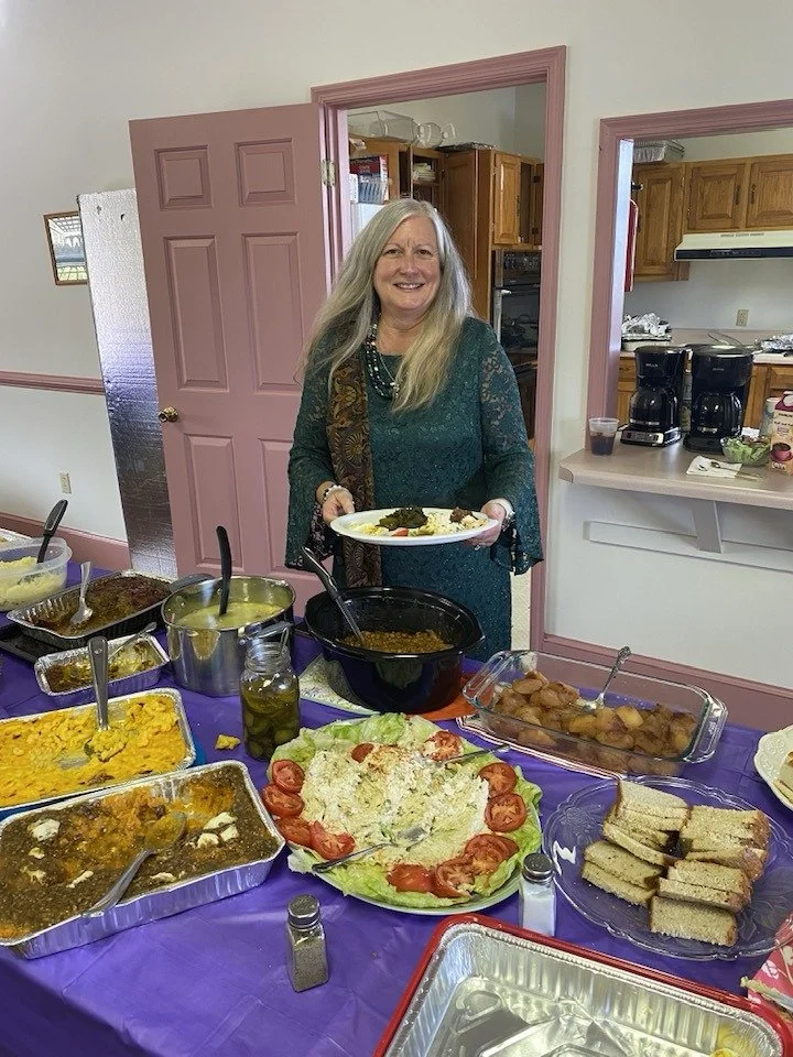 A woman with long blonde hair in a green lace dress smiling and holding a plate of food at a buffet table with various dishes, in a room with pink trim and a kitchen in the background.