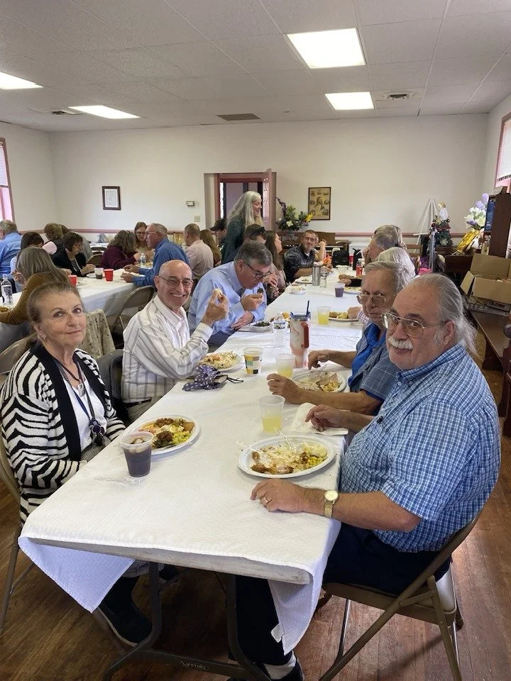 A group of older adults sitting at a long table in a community room, enjoying a meal together. The room has white walls, fluorescent lighting, and a few decorations. The table is covered with a white tablecloth and has plates of food, drinks, and condiments.