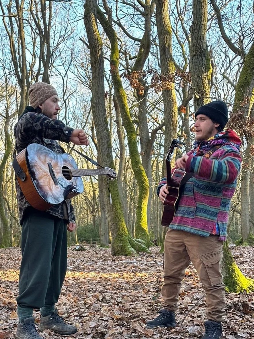 Two men playing acoustic guitars in a wooded area during autumn. The man on the left is wearing a brown beanie, black jacket, and green pants. The man on the right is wearing a black beanie, colorful patterned jacket, and tan pants.