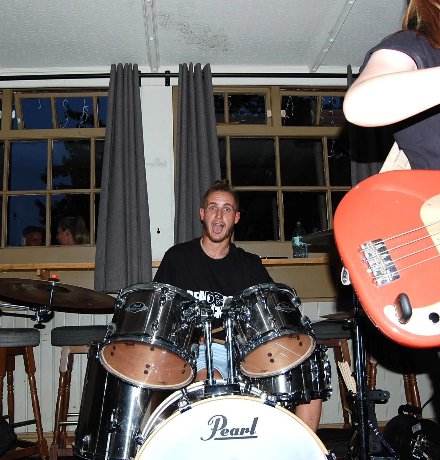 A person sitting behind a drum set, making a funny face, with another person holding a guitar partially visible in the foreground, in an indoor setting with windows and curtains in the background.