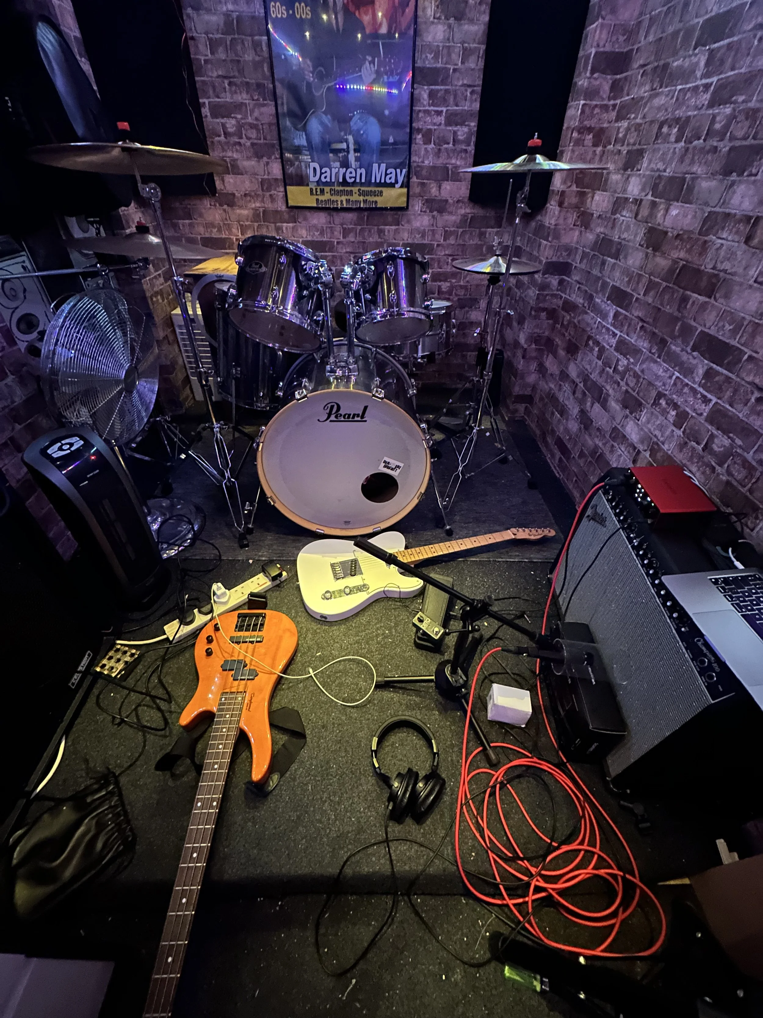 Concert stage setup with a drum set, two electric guitars (one orange and one white), a microphone on a stand, headphones, an amplifier, and other music equipment in a brick-walled room.