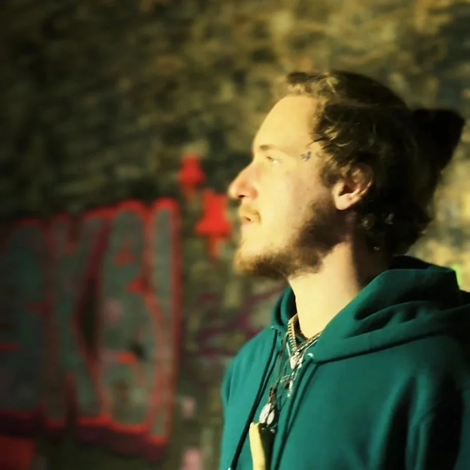 A young man with a beard and long hair tied back, wearing a green hoodie, standing in front of a graffiti-covered wall, looking to the right.