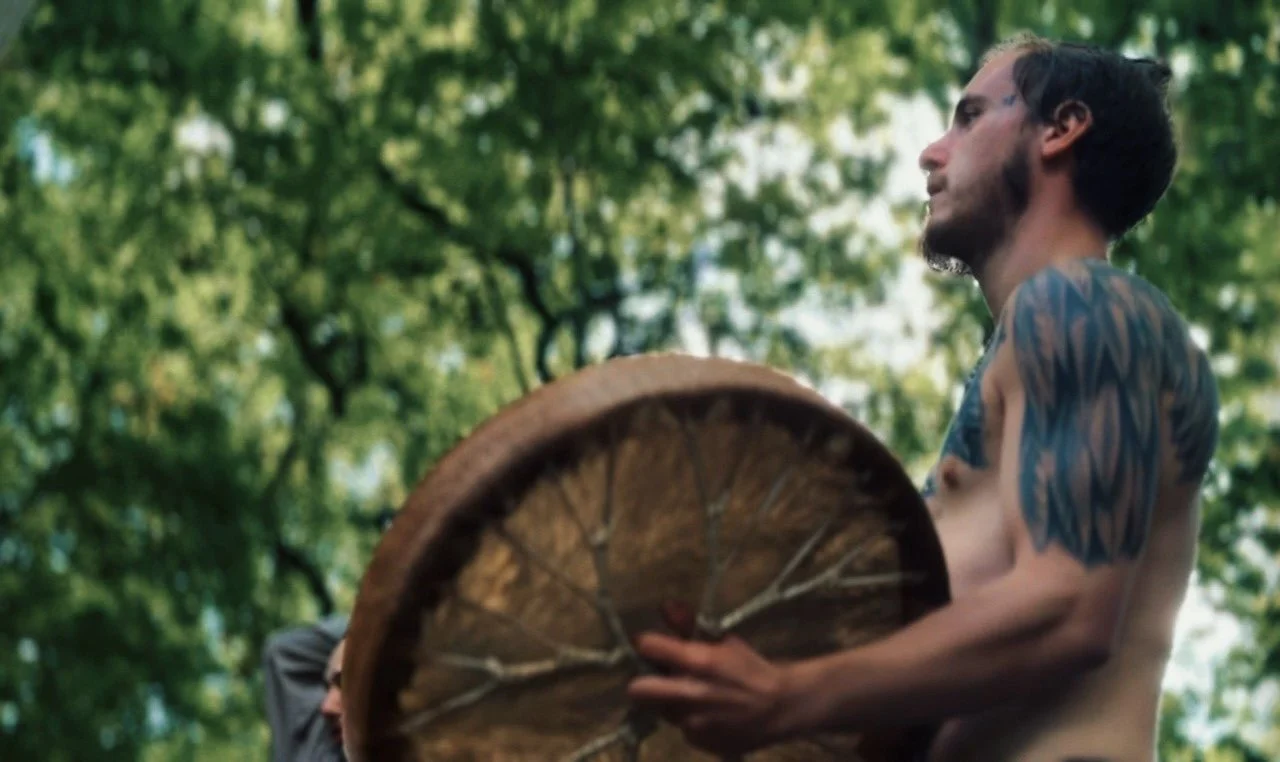 A shirtless man with tattoos on his arm and a side profile, playing a shamanic drum, standing outdoors with green trees in the background.