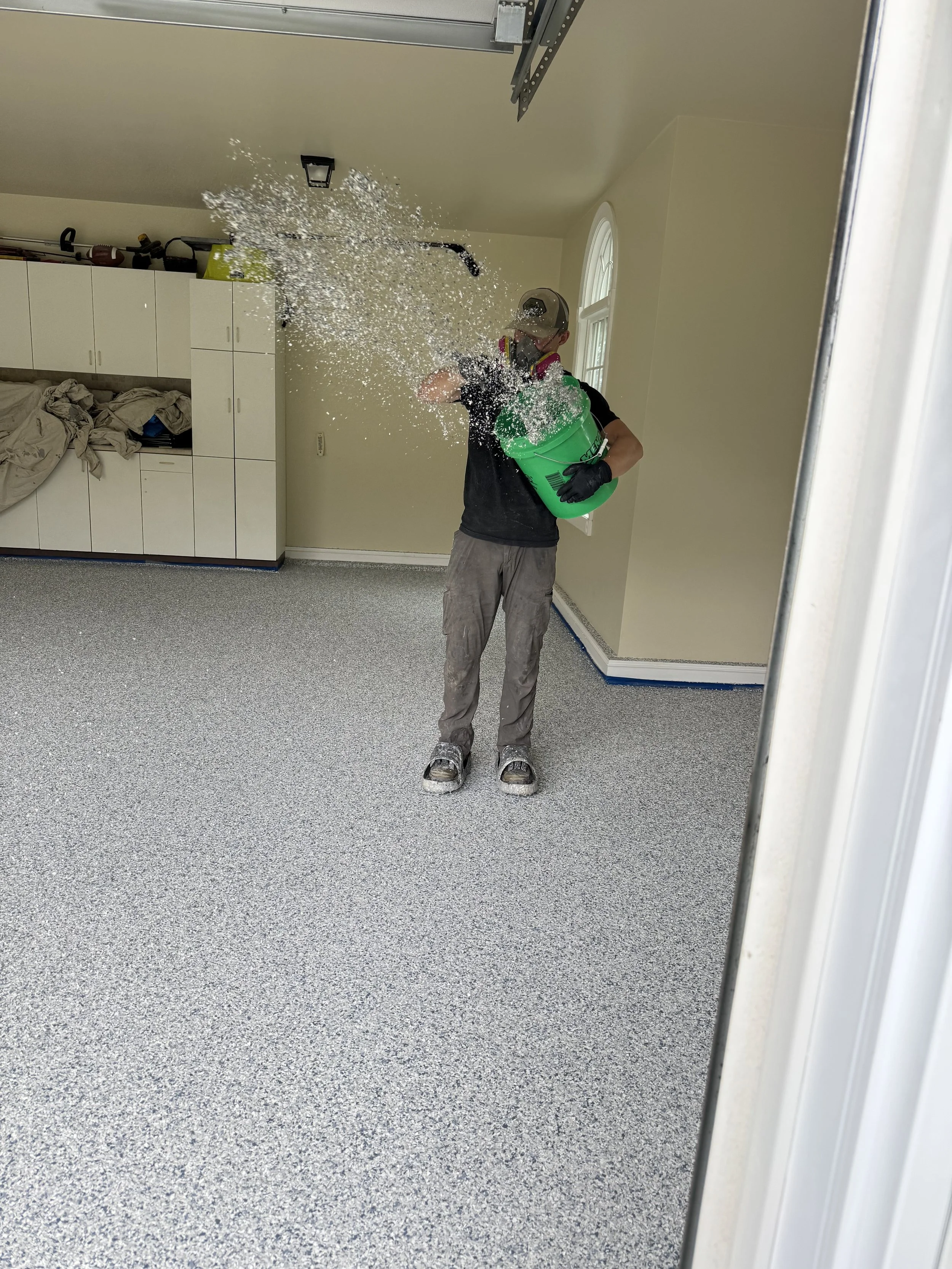 A person wearing a cap, face mask, black gloves, and black shorts is standing on gray carpet, holding a green bucket, and spilling water foam while cleaning a room with white cabinets and a gray ceiling.