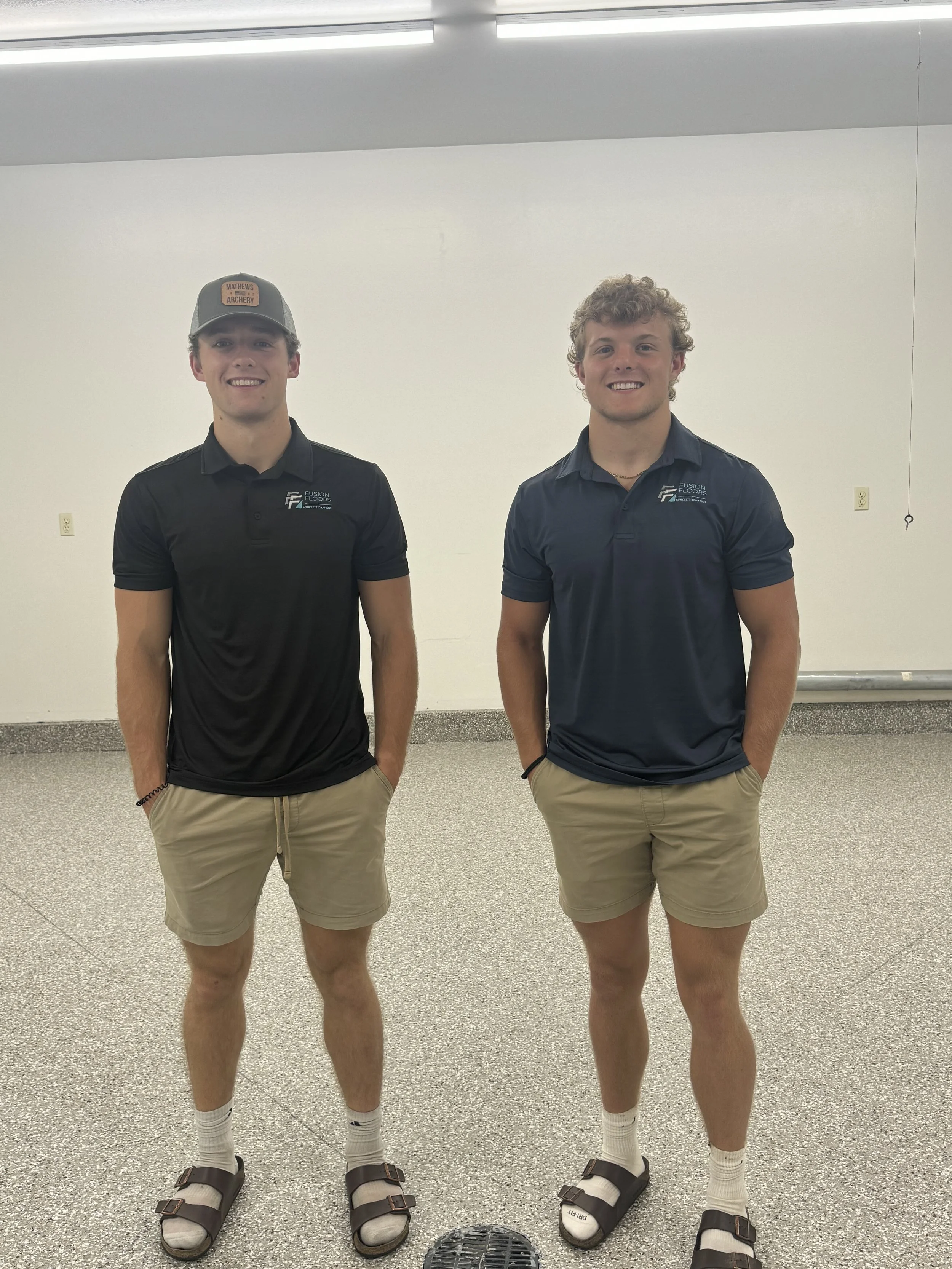 Two young men standing in a room with a white wall background, both wearing dark polo shirts with a logo, beige shorts, white socks, and sandals. One is wearing a cap, and they are both smiling with their hands in their pockets.