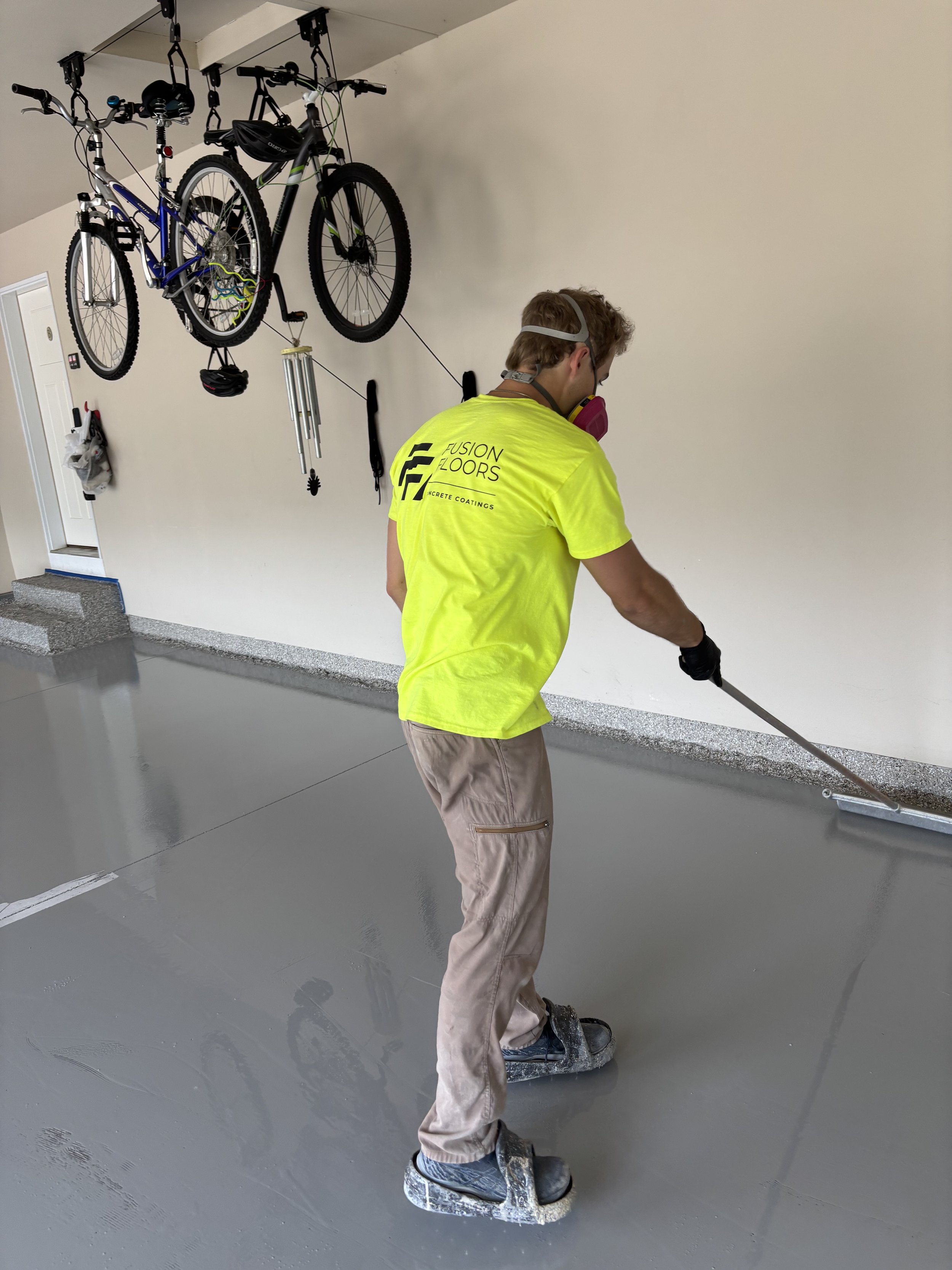 A person wearing a yellow shirt, beige pants, and a face mask is using a long-handled tool to spread or smooth fresh concrete on a floor in an indoor space. Two bicycles are hanging from the ceiling in the background.