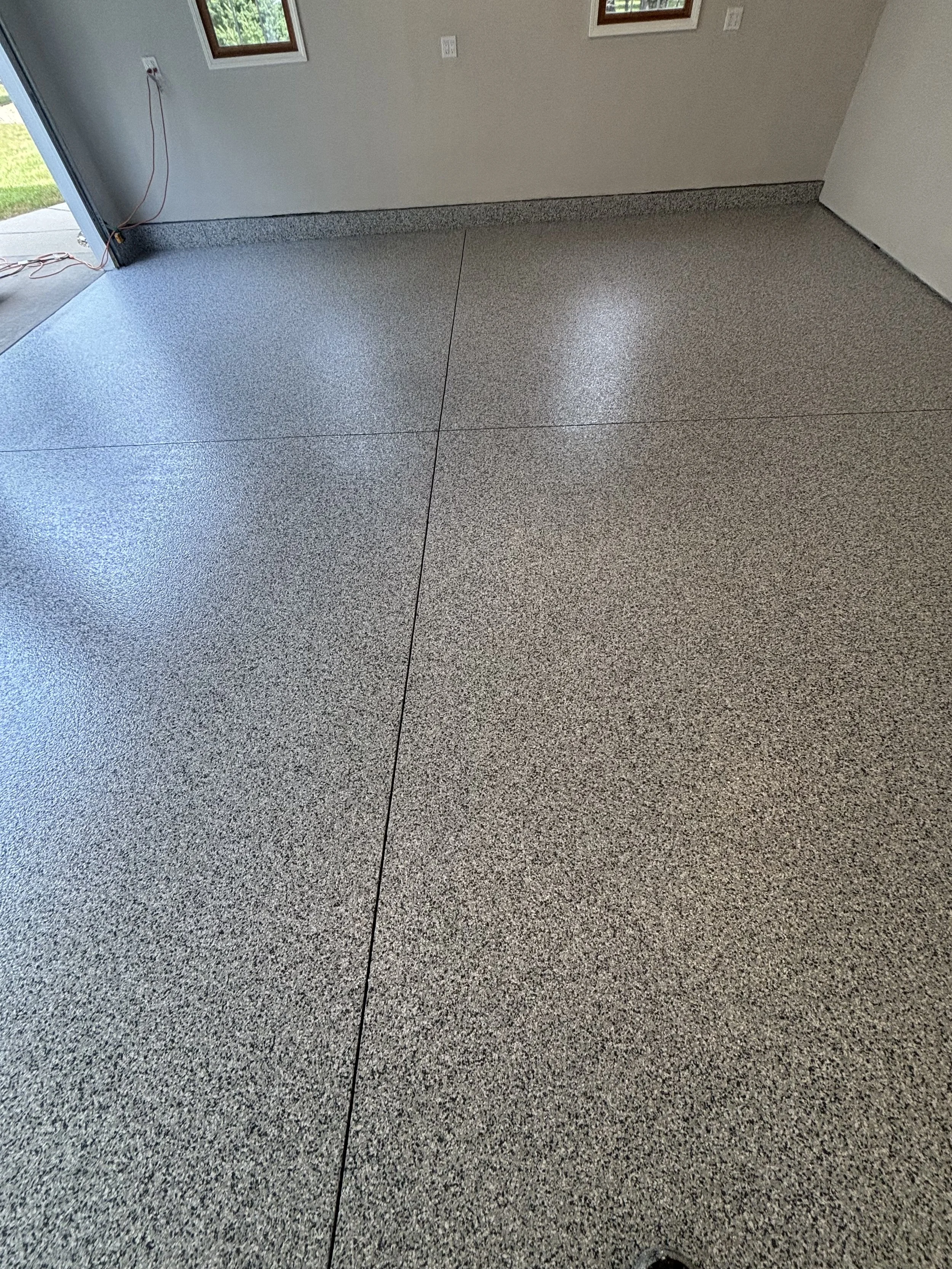 Empty garage interior with speckled gray epoxy floor, white walls, and two small windows. Some electrical cords are hanging near an open garage door.