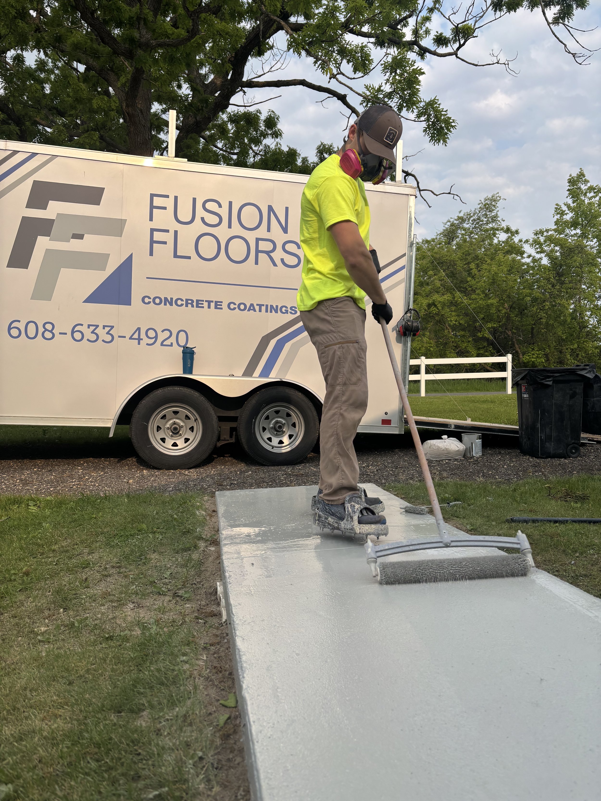 A worker wearing a bright yellow shirt, gray pants, a brown cap, and pink ear protection is operating a concrete grinder on a concrete surface outside. There is a large white trailer with company branding and a phone number in the background, along w
