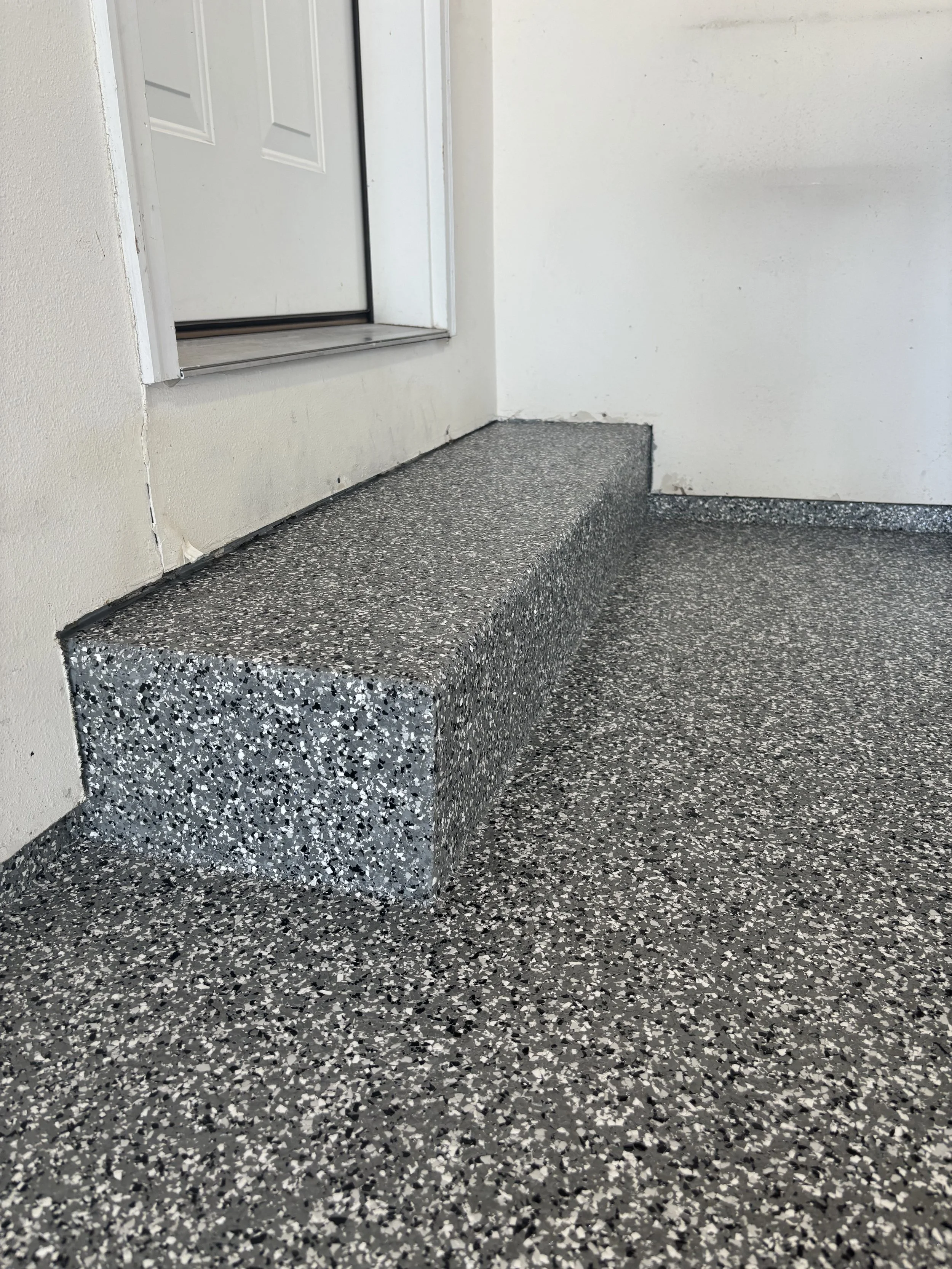 Close-up of a building floor with a terrazzo pattern and a step leading up to a white door with a silver threshold.