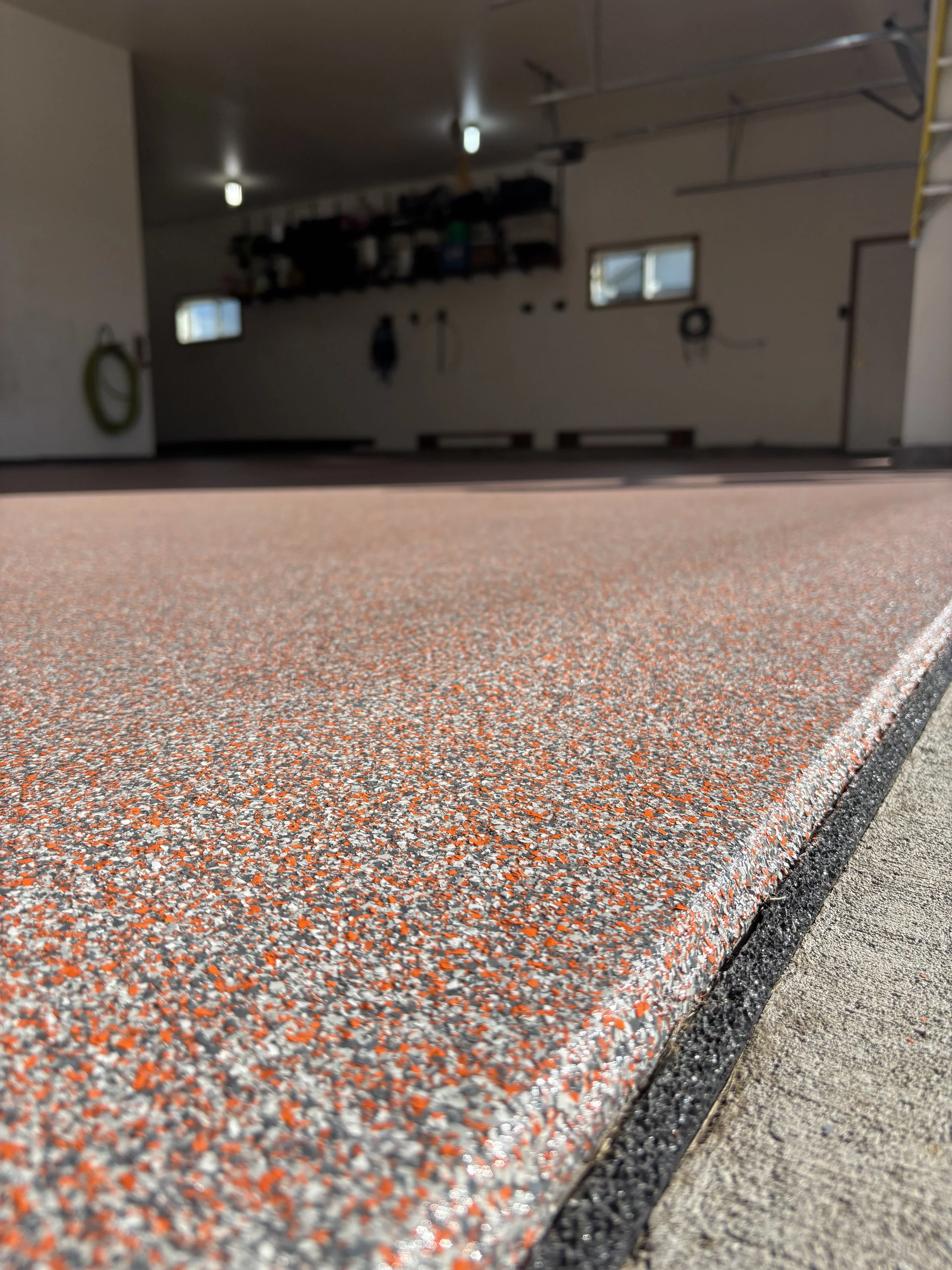 Close-up of a speckled red, gray, and white epoxy garage floor with black edging and a concrete driveway border.