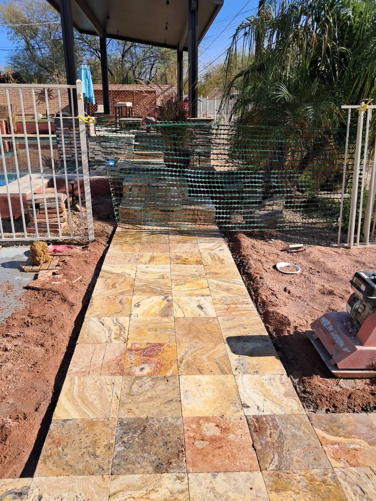 A newly constructed stone patio with a pathway surrounded by dirt in a backyard. The patio is bordered by a white metal fence, and there is a potted plant on a ledge in the background under a covered porch. In the background, there are trees, a brick