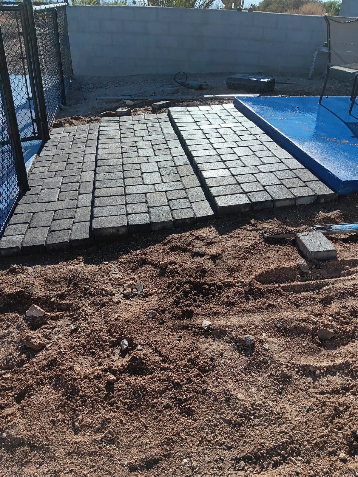 Under construction backyard with newly laid stone pavers, a blue painted platform, tools, and a metal fence.