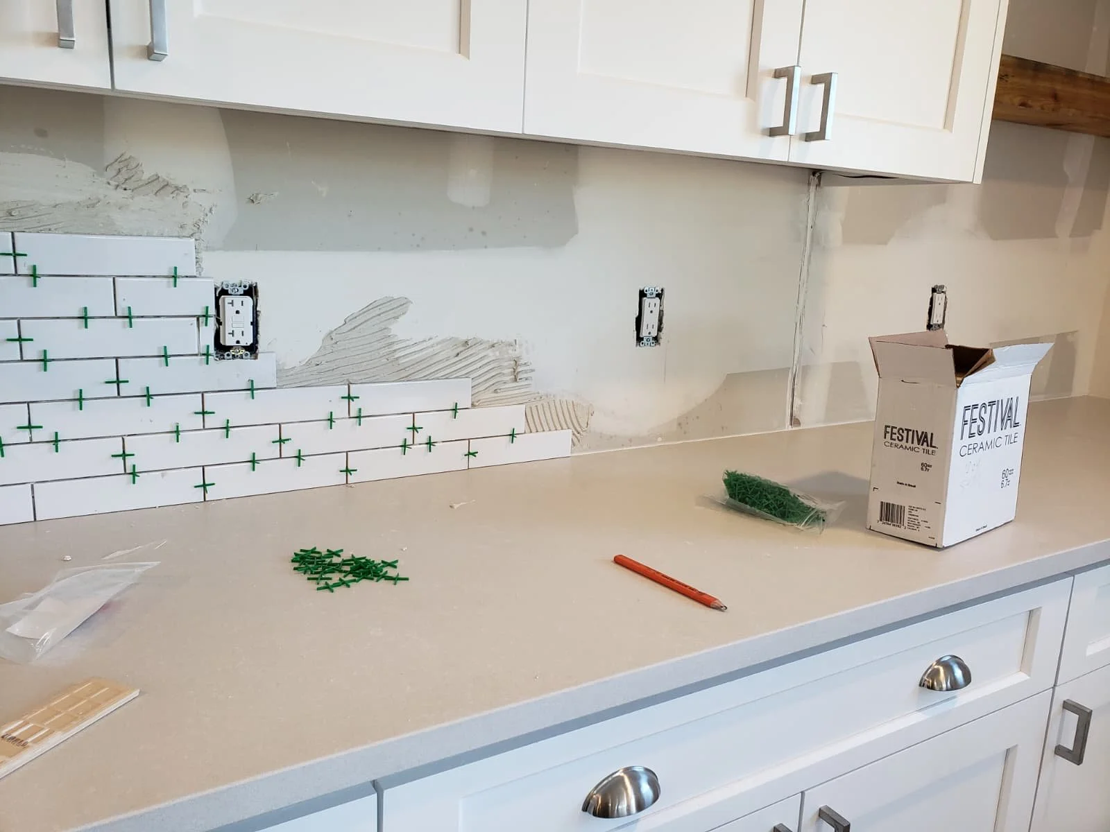 Kitchen countertop with tiles, a box of ceramic tiles, a green brush, and a red pen, with white cabinets above and a partially tiled wall.