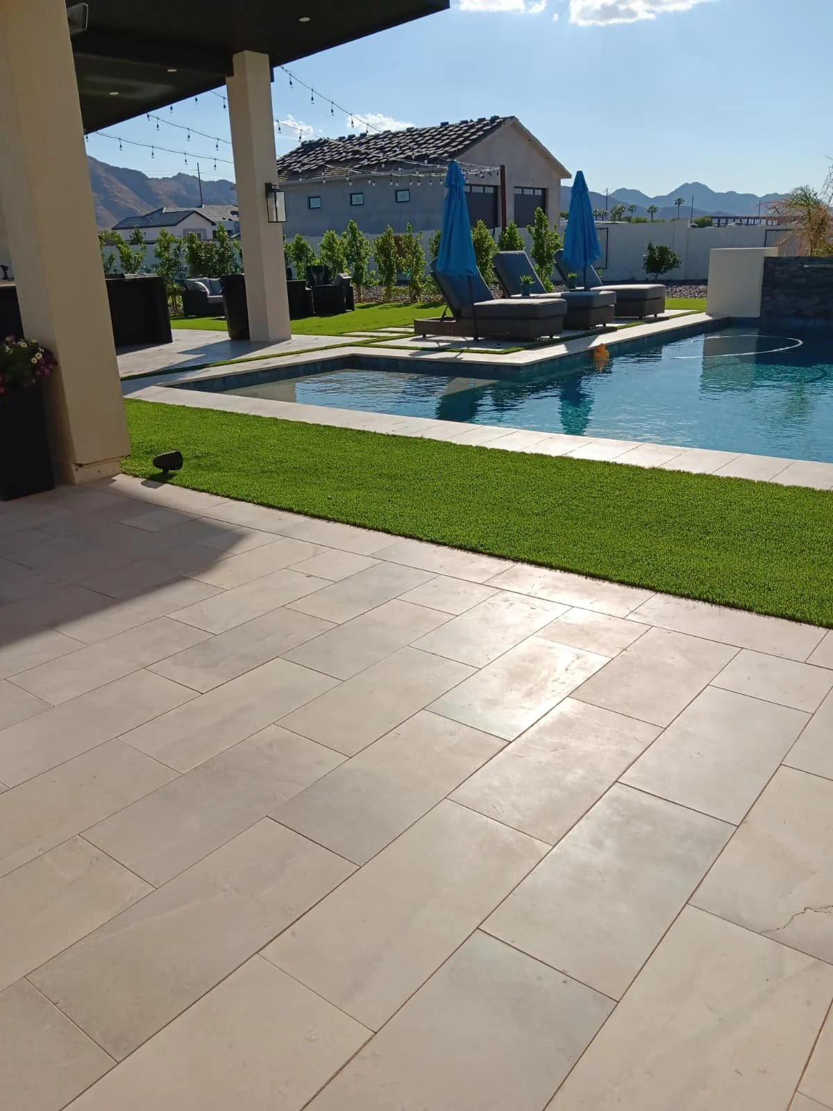 Image of a backyard with a swimming pool, lounge chairs with blue umbrellas, a small grassy area, and distant mountains under a clear sky.