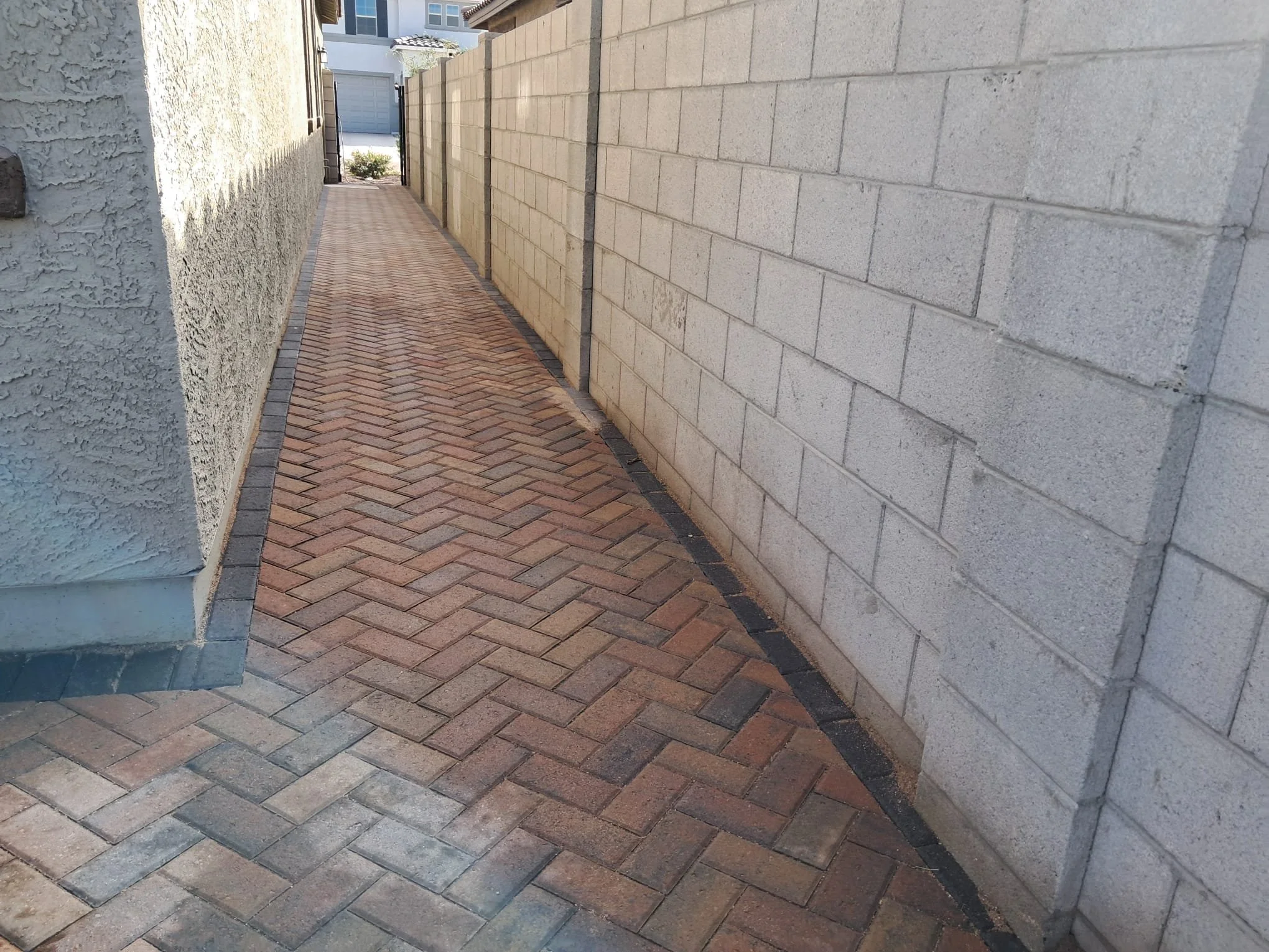 Narrow brick pathway between two walls, one stucco and one brick, leading to a gate or opening at the end.