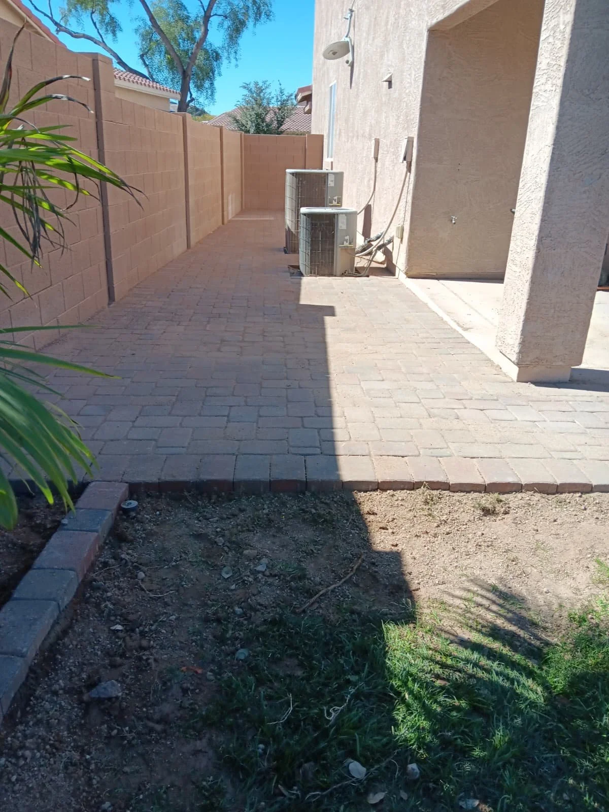 Backyard patio with brick pavers, beige stucco walls, and two air conditioning units. There's a wooden fence at the end, a tree in the background, and shadows cast on the patio.