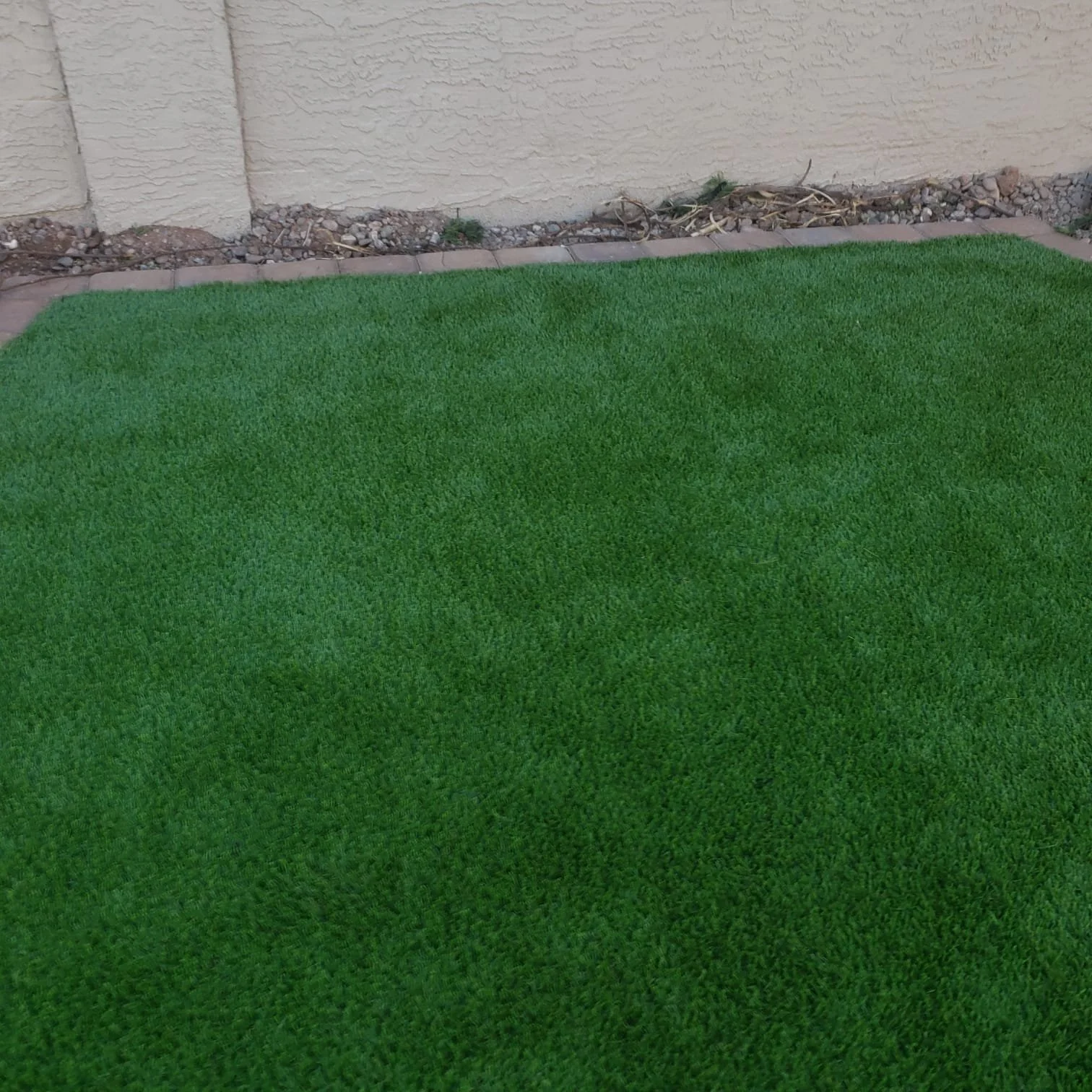 Artificial green turf grass outside in front of a beige wall with small rocks along the edge.
