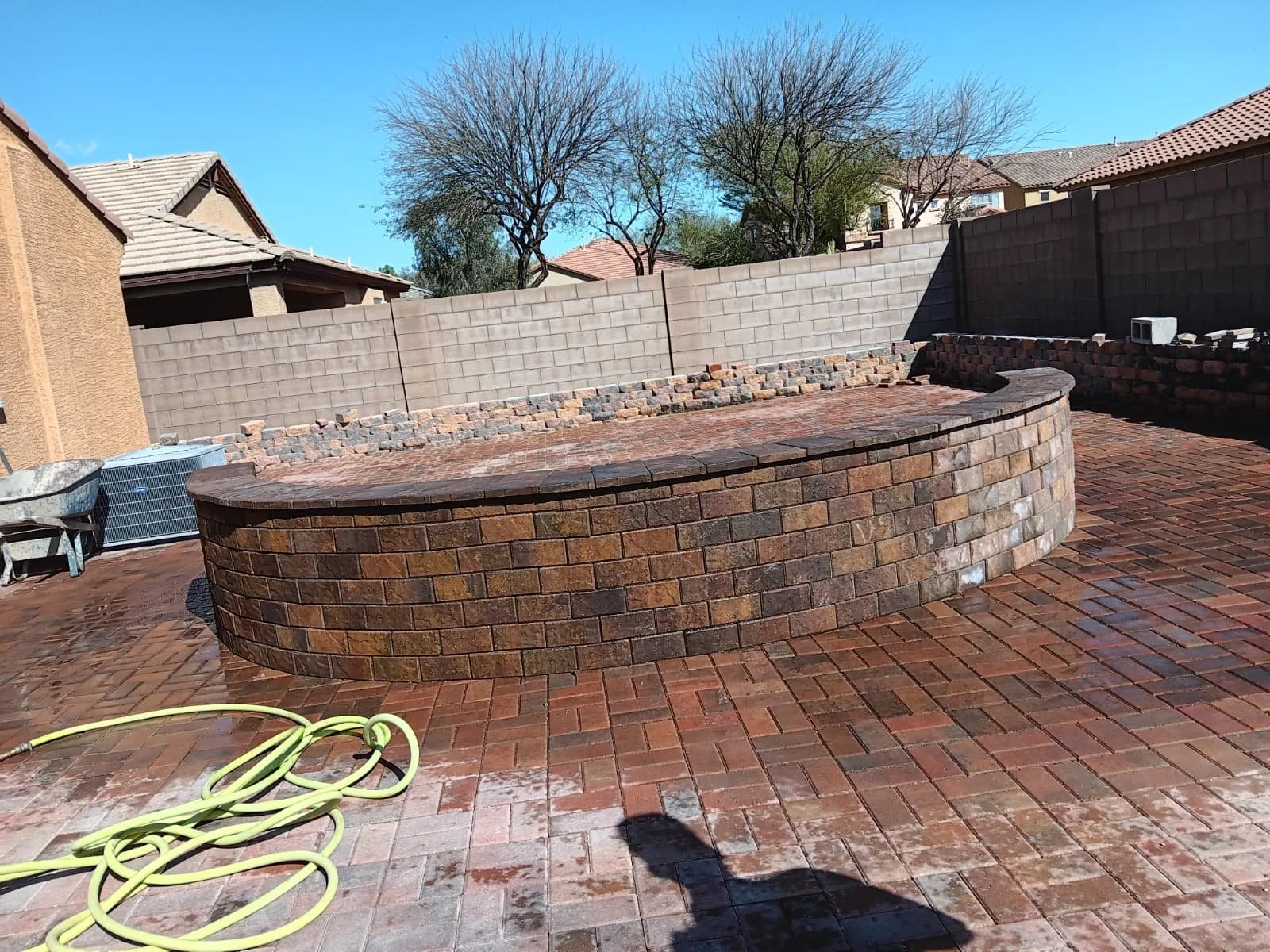 Backyard with a raised brick patio, a curved brick wall, and a tiled floor, surrounded by a privacy fence with neighboring houses and trees in the background.