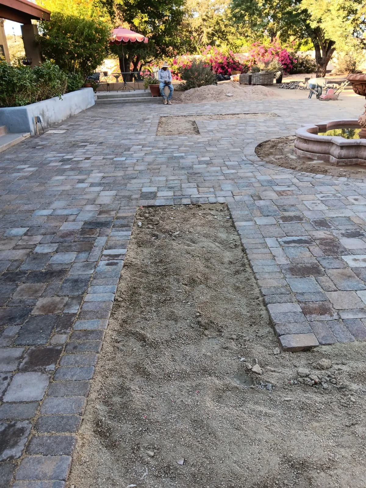 A backyard with a brick patio under construction, with some areas of dirt and paving stones missing. There are plants, trees, a fountain, and a person sitting and reading under a red umbrella in the background.