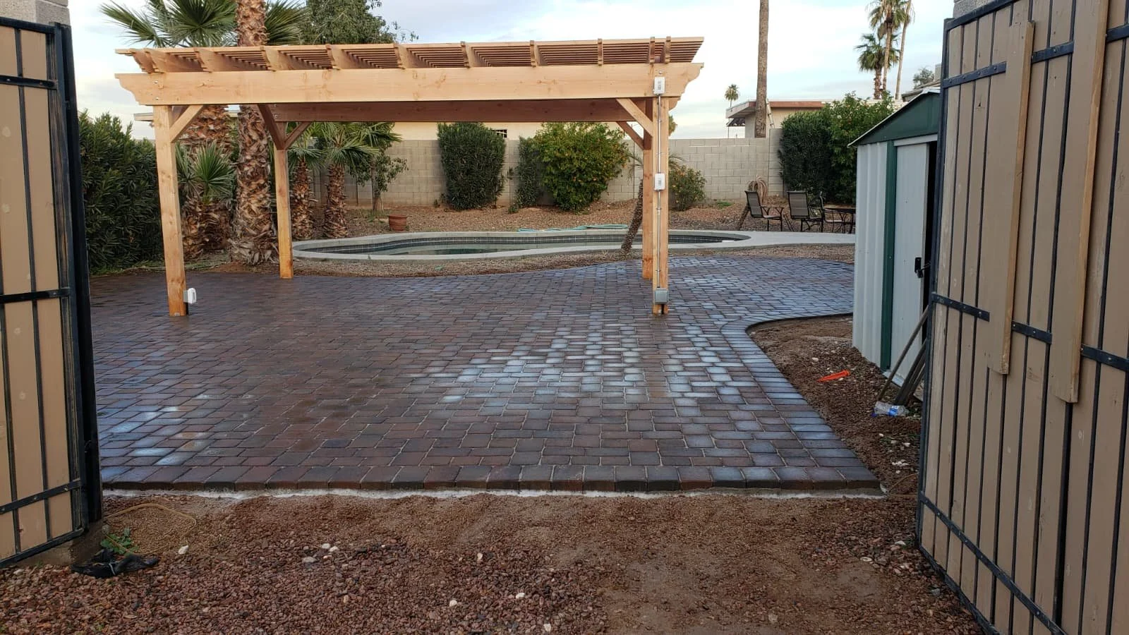 View of a backyard with newly installed brick paver patio, metal pergola, small pool, and outdoor seating area, surrounded by desert landscaping and a wooden fence.