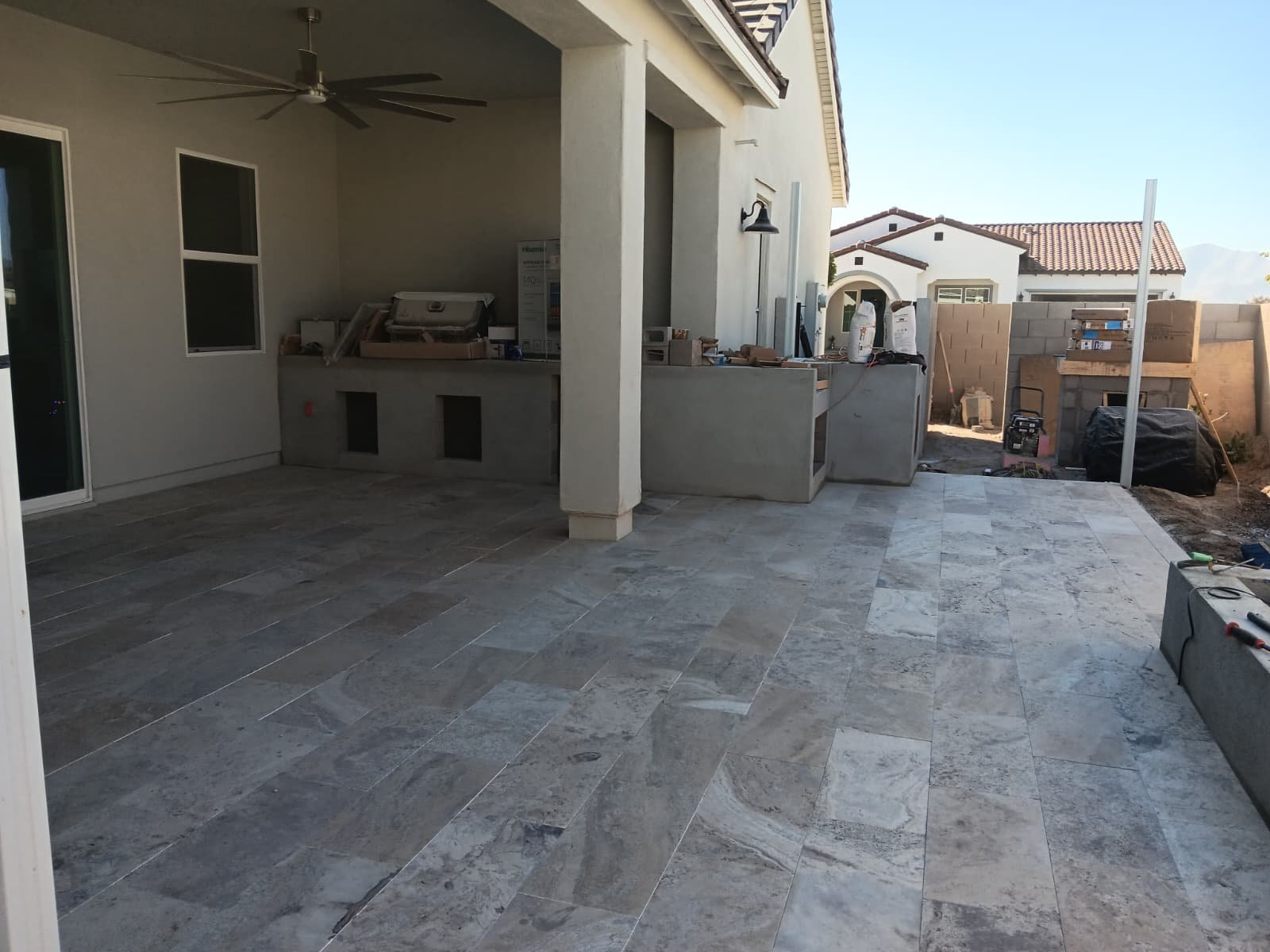 Back patio under construction with stone tile flooring, partially built outdoor kitchen area with concrete counters, and unfinished yard with stacks of bricks and construction materials.