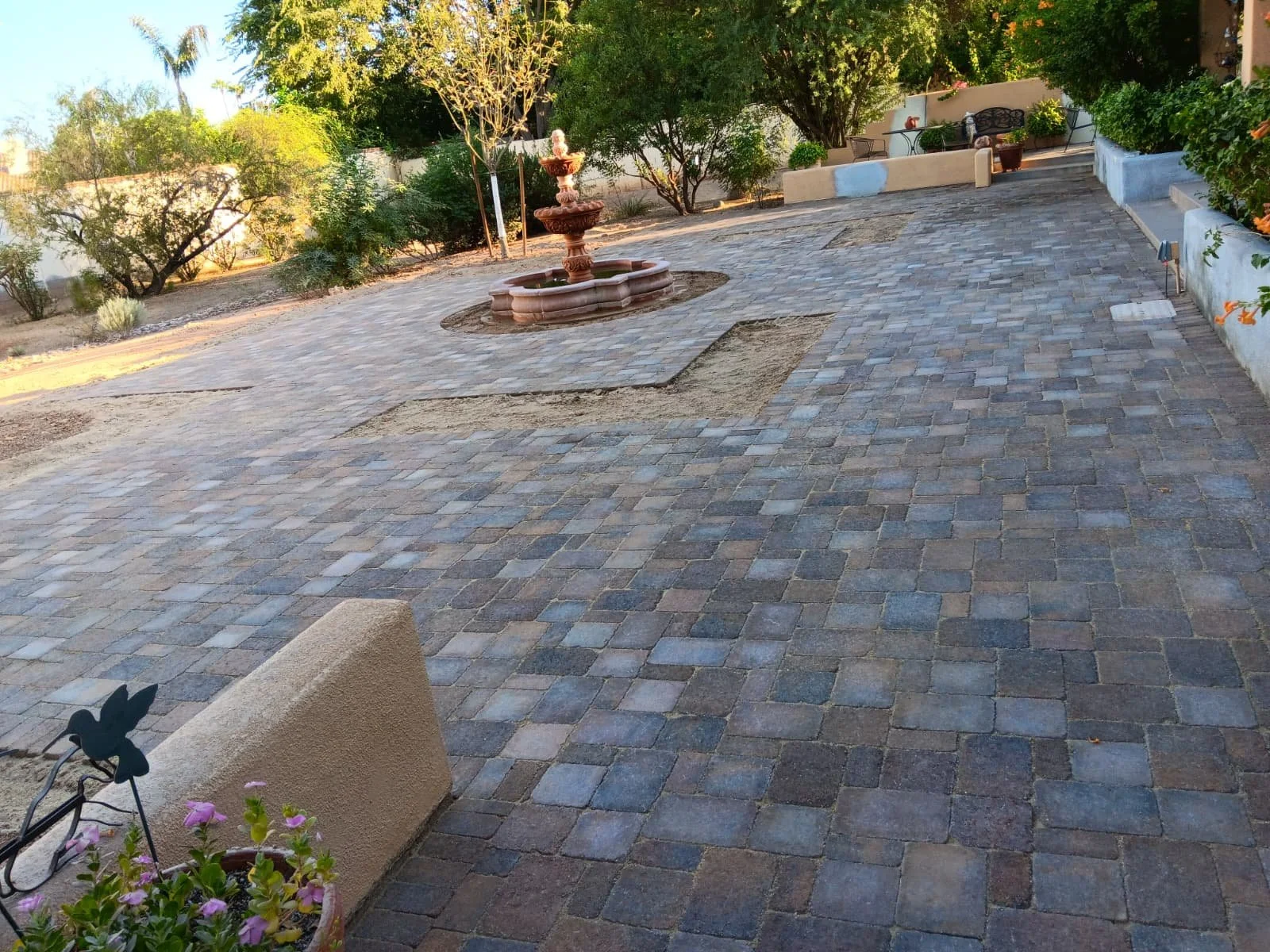 A paved courtyard with a decorative fountain at the center, surrounded by trees and garden furniture, with some potted plants in the foreground.