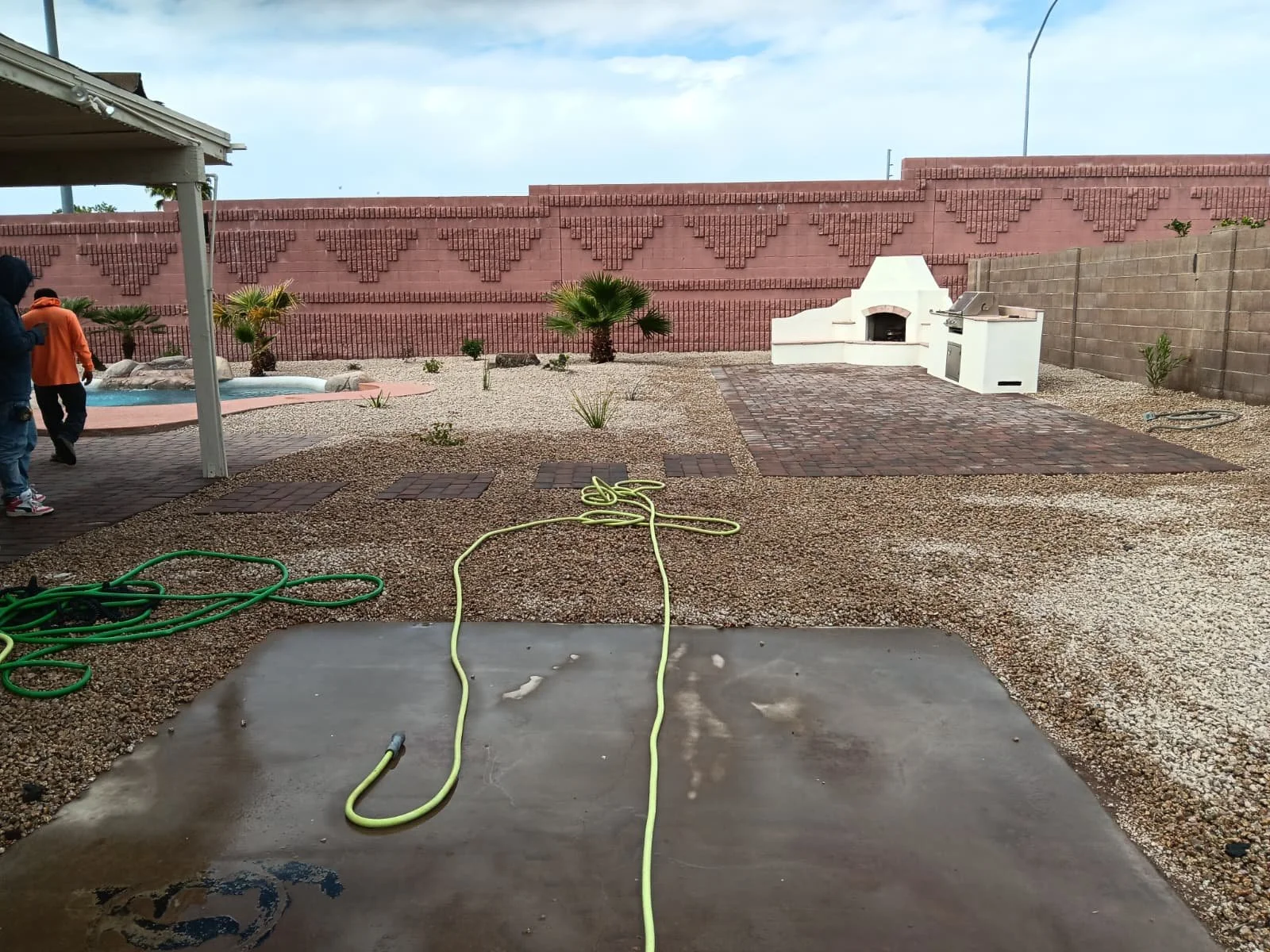 Backyard with a brick patio, a small fireplace, a white outdoor kitchen, gravel ground, palm trees, and two people standing near the patio.