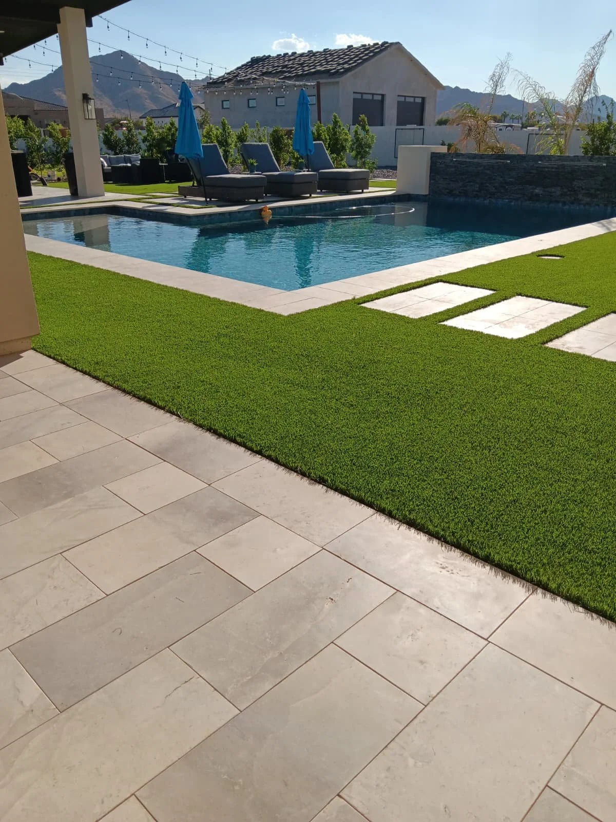 Backyard with a swimming pool, lounge chairs with blue umbrellas, artificial grass, and tiled patio with mountain view in the background.